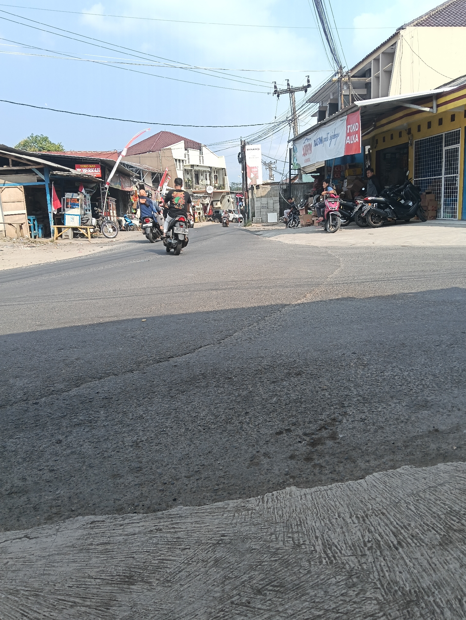 In Puspahiang, Indonesia, a street scene unfolds under a clear sky. A road, partially paved with asphalt and concrete, curves into the distance. On the right, a row of buildings features shops with yellow and white facades. One shop displays a red banner with white text reading "TOKO ALKA." Several motorcycles are parked in front of these shops, some with riders. On the left side of the road, a smaller, more rustic structure with a tin roof is visible, also with parked motorcycles. In the middle of the road, two individuals are riding motorcycles, one in the foreground with a black shirt and the other behind with a blue shirt. Further down the road, other vehicles and people are scattered, suggesting active commerce and traffic. Indonesian flags are displayed on poles near the buildings on the left. The environment appears to be a typical urban or semi-urban area, characterized by a mix of modern and traditional structures and the presence of motor vehicles.