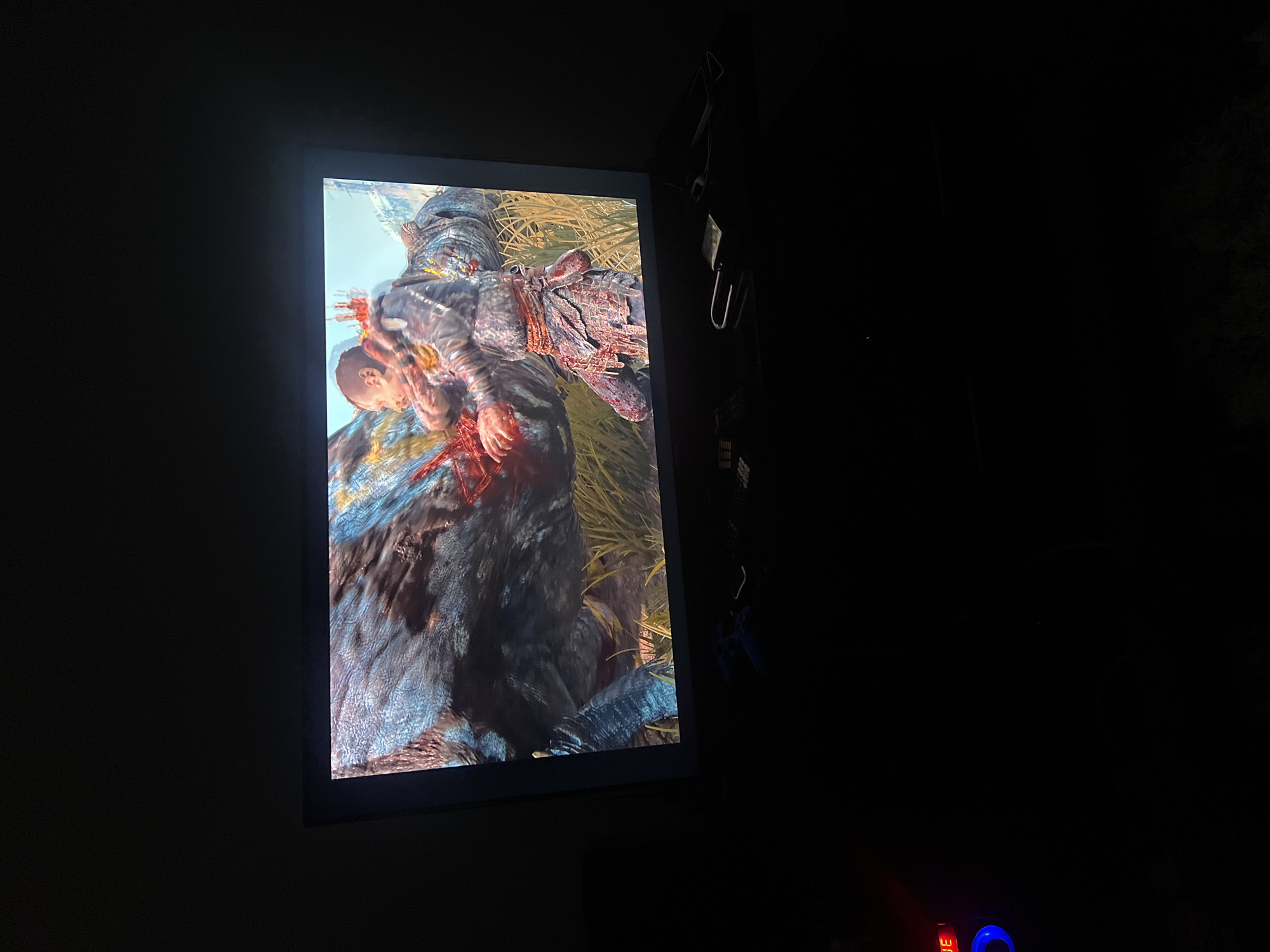 The image displays a television screen in a dimly lit room, showing a scene from a video game. On the screen, a young male character with short hair, wearing a fur-lined tunic and patterned leggings, appears to be in combat with a large, dark-skinned creature with textured skin and gold accents. The character is holding his arms up, and there is visible red on his hands and the creature's body, suggesting violence or injury. The background of the game scene features tall, dry grass and a bright sky. Below the television, several objects are visible on a dark surface, including a blue video game controller, a remote control, and a padlock. A red digital display with the letters "JE" is also visible on the far left. The overall environment is dark, with the television screen being the primary source of light. There is no indication of the location being in Jos, Nigeria.