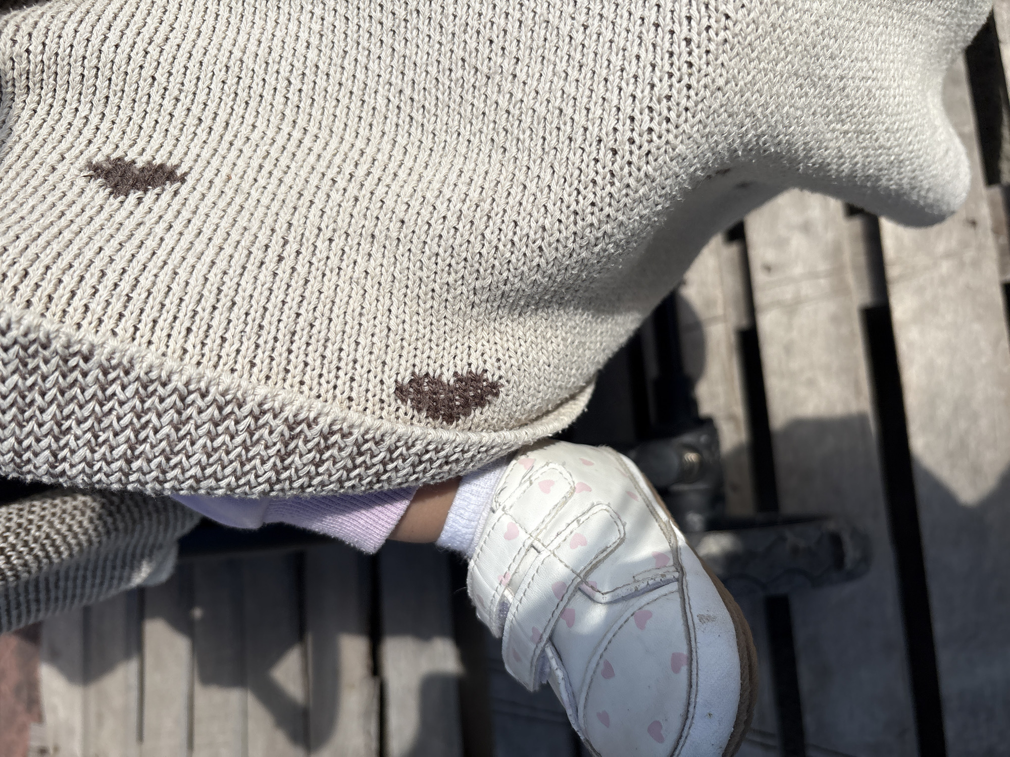 A close-up, high-angle shot depicts the torso and left leg of a child wearing a light-colored knit sweater with dark brown heart patterns. The child wears a white sock and a white sneaker with pink heart decorations. The child is sitting on a wooden surface, possibly a deck or steps. A piece of light purple clothing is visible between the sweater and the child's sock. The location is Ембаево, Russia.