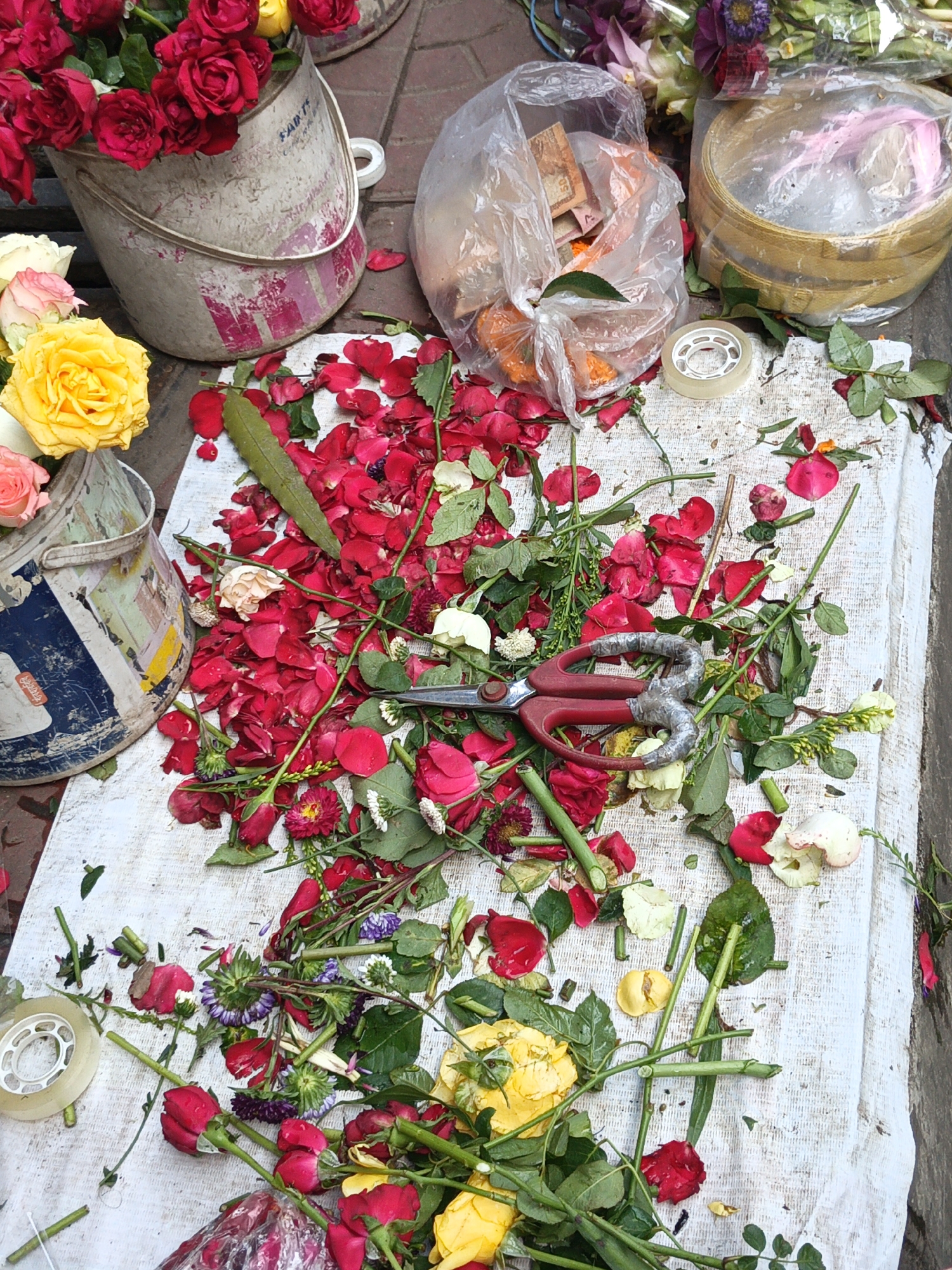 The image shows a street-level flower stall in Dhaka, Bangladesh. On a white cloth spread on the ground are scattered rose petals, stems, and whole flowers (red, yellow, and white). A pair of red-handled scissors lies amidst the flower debris. Two buckets, one containing red roses and another with yellow and pink roses, are positioned on the upper left. A clear plastic bag holding orange marigolds and a bamboo steamer-like container are on the upper right. A roll of transparent tape is also visible. The scene depicts the remnants of flower arrangement activities.
