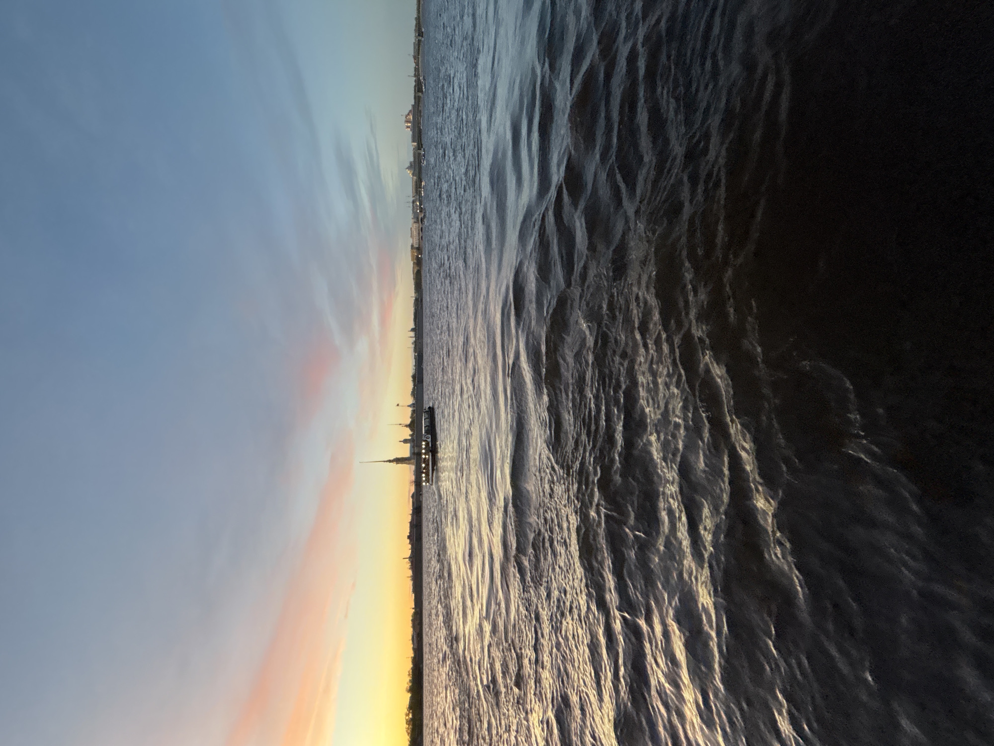 The image depicts a waterfront scene at sunset in Saint Petersburg, Russia. The sky displays hues of blue, orange, and yellow. The silhouette of the Peter and Paul Fortress, with its iconic spire, is visible in the distance. A body of water, likely the Neva River, dominates the foreground. The river's surface is textured with ripples and waves, reflecting the colors of the sky. A boat with illuminated lights is docked near the shore. The cityscape of Saint Petersburg stretches along the horizon, with buildings and structures silhouetted against the fading light. The right side of the image is dark.
