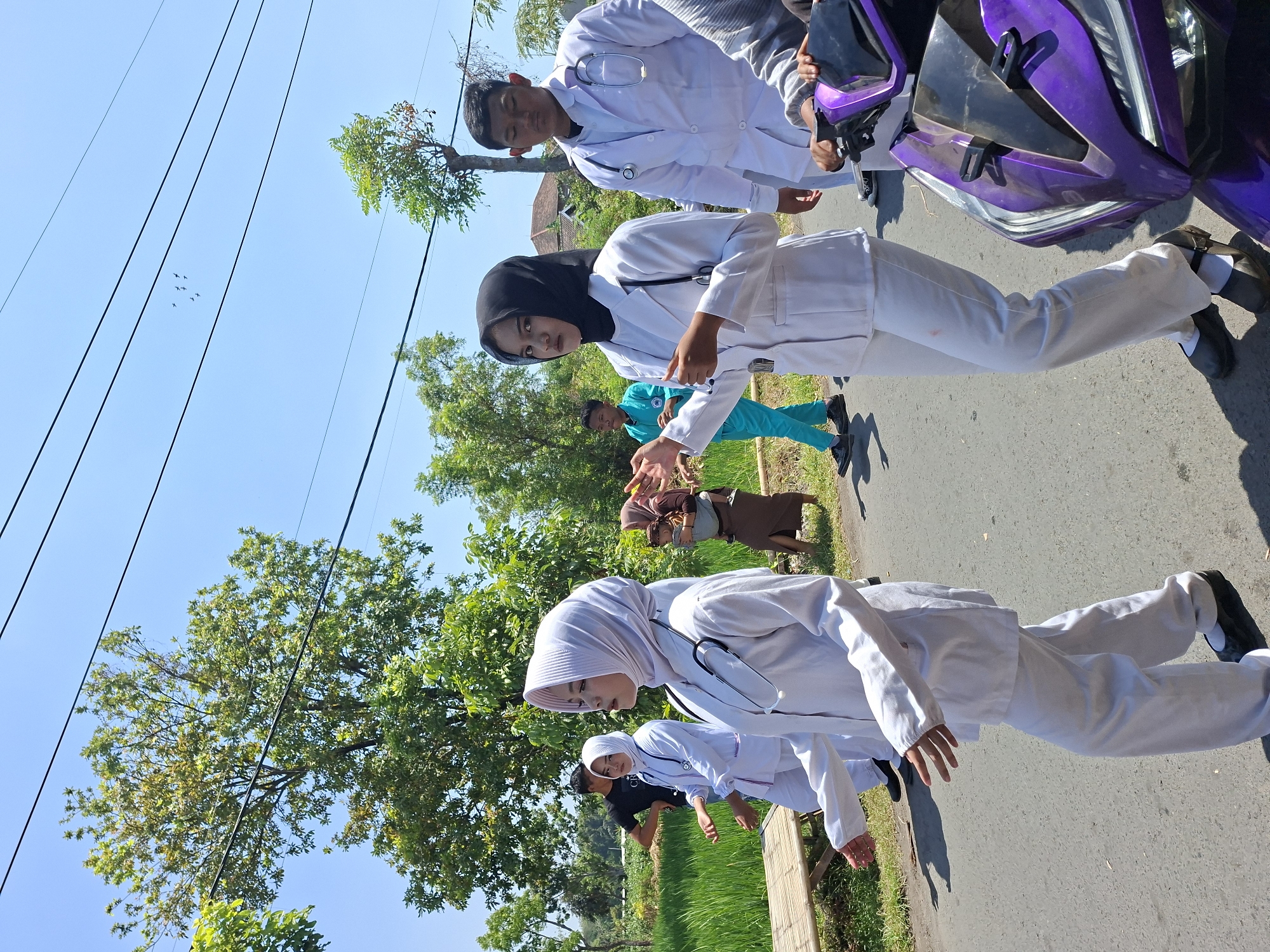 In Tongaswetan, Indonesia, a group of individuals in white medical attire walks along a paved road under a clear blue sky. The foreground features a purple scooter. To the left, a person in a black t-shirt observes. Behind them, two women in white lab coats and hijabs, one in a light pink hijab and the other in a black hijab, are prominent. The woman in the pink hijab is in the foreground, her arm outstretched. The woman in the black hijab stands slightly behind her, also with an outstretched arm. Further back, a woman carries a baby wrapped in a cloth carrier. Several trees with lush green foliage dot the background, along with a rice paddy field on the left side of the road. The overall scene depicts a daytime outdoor gathering in a rural or semi-rural Indonesian setting.
