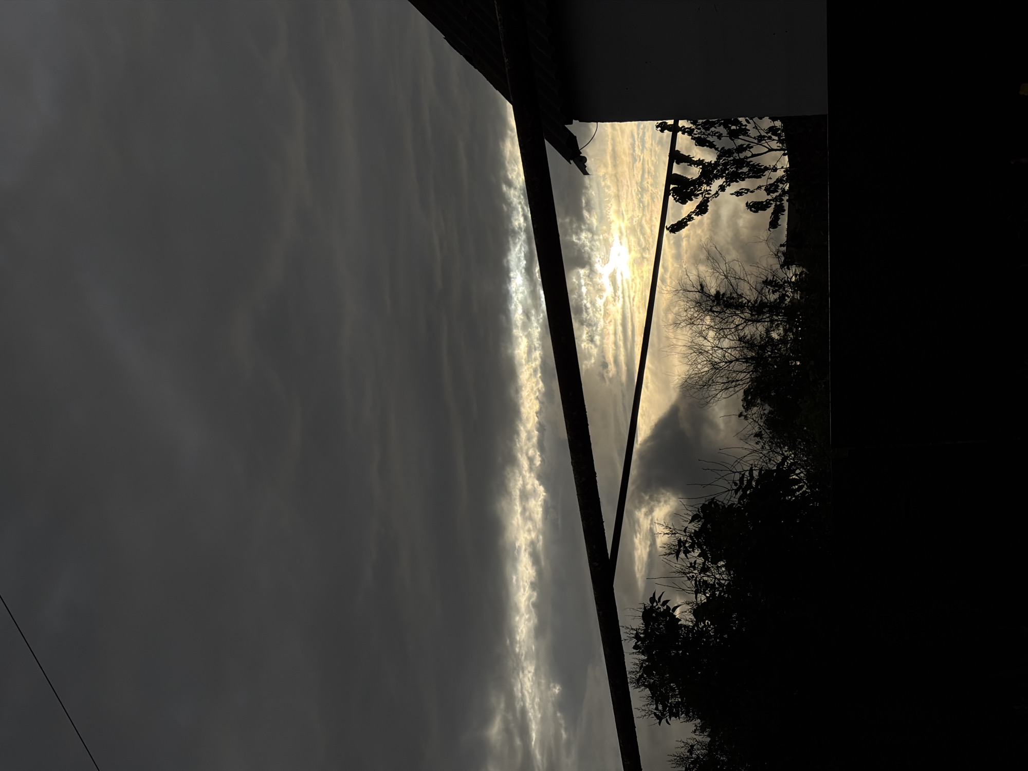 The image presents a view of the sky and elements of an urban scene, likely taken in Novopokrovskaya, Russia. Overcast conditions dominate the sky, with a long, thin band of lighter clouds suggesting the location of the sun. A building edge and metal support structures are visible at the top of the frame, transitioning into silhouette along the right edge. Trees with dense foliage are also silhouetted against the brighter sky. The lower right of the image is predominantly dark, suggesting shadow and obscuring details. A thin power line stretches across the lower left corner. No people are visible.