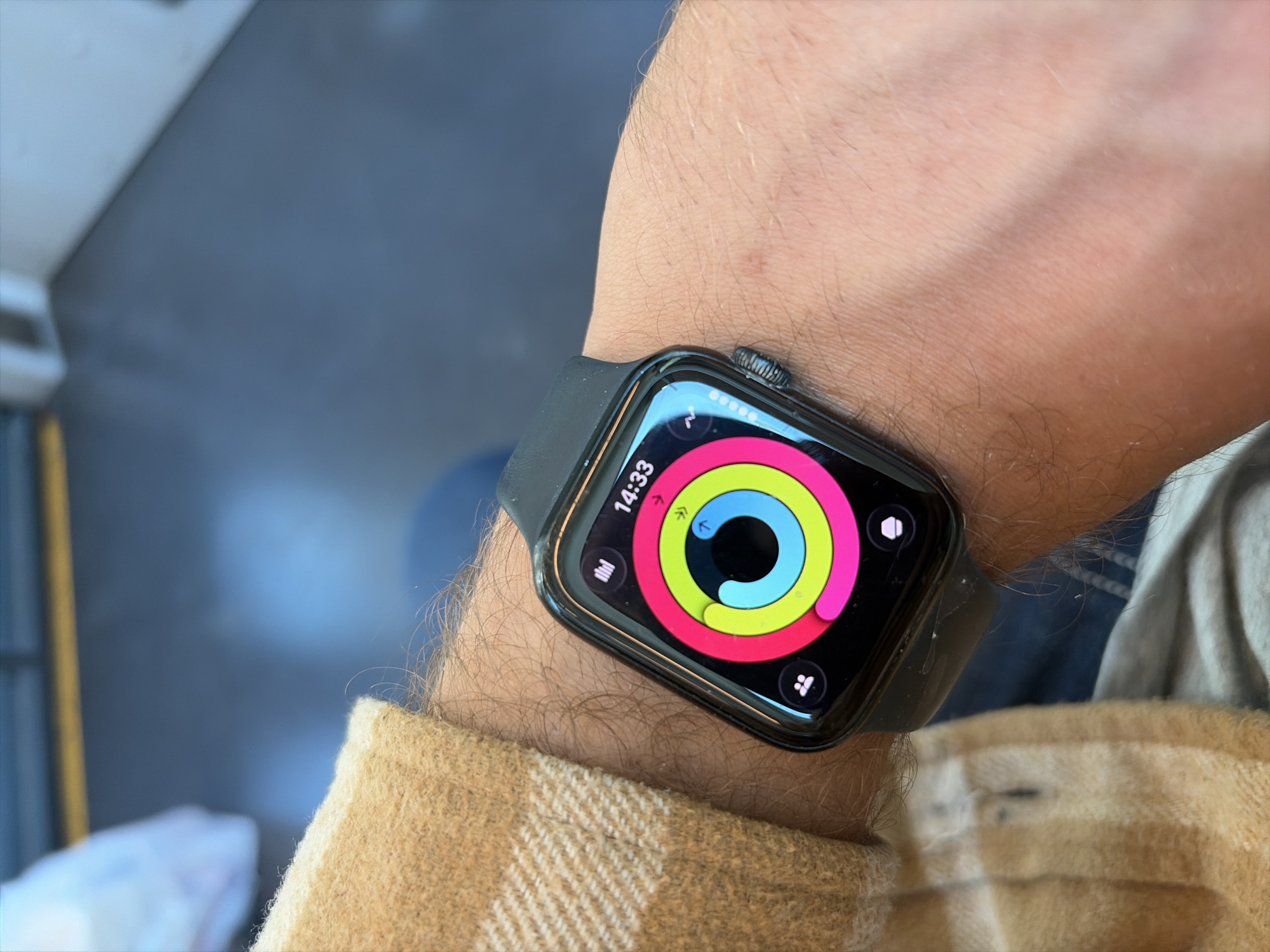 A close-up shot shows a person's wrist with an Apple Watch. The watch face displays the activity rings and the time 14:33. The wrist is hairy, and the person is wearing a tan-colored shirt. The background is out of focus, but it appears to be an indoor setting with gray and blue tones. There is no information to confirm the location is in the city of Jerusalem, Israel.