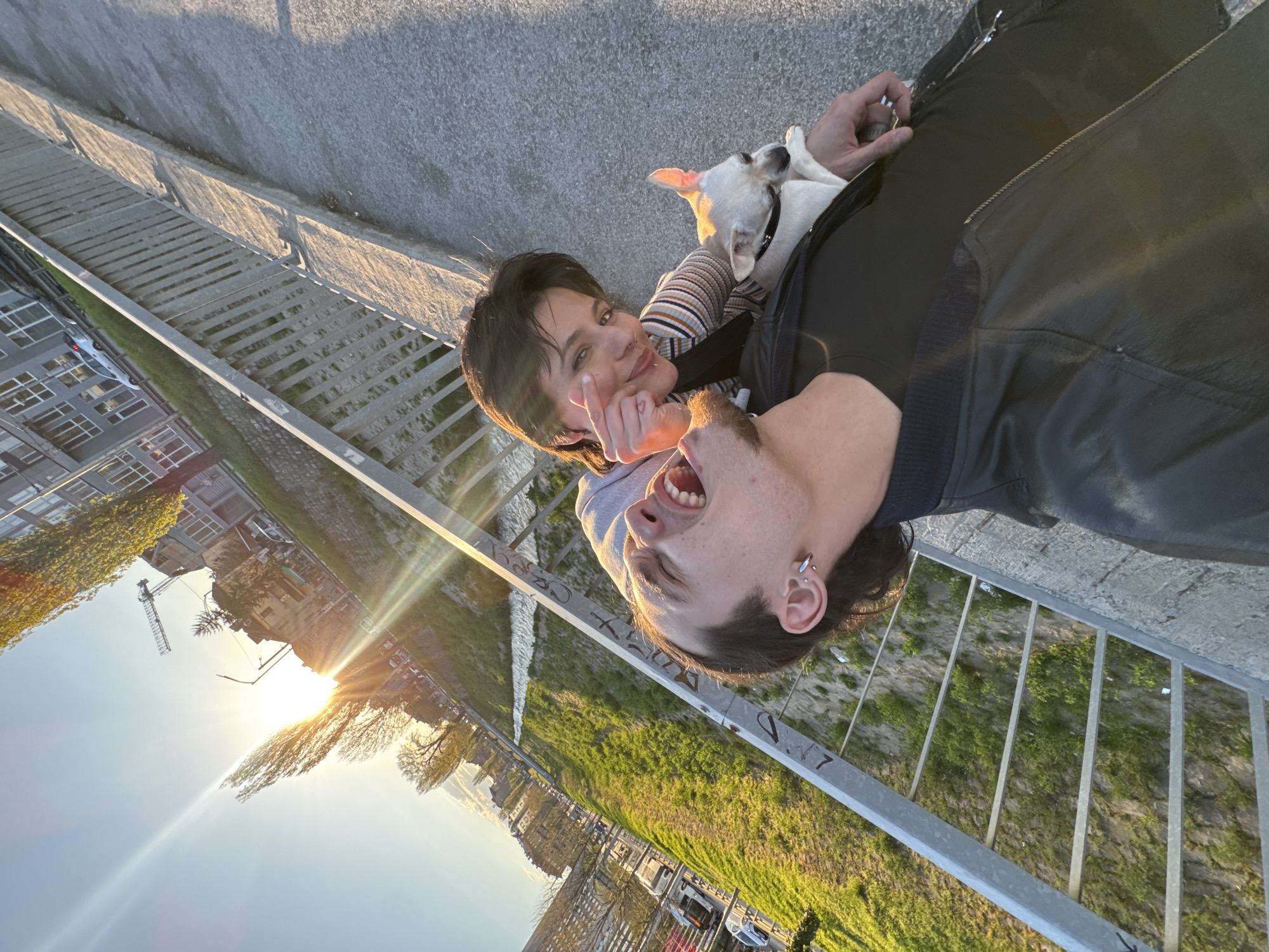 A sunlit, off-kilter, eye-level shot depicts a man and woman with a small white dog on a bridge in what appears to be Sofia, Bulgaria. The man, wearing a black leather jacket, is turned towards the camera with his mouth open. The woman, wearing a striped top and a grey sweatshirt, is next to him, smiling and holding up two fingers. A small, white dog is nestled between them. The bridge has a silver metal railing, and below it, green vegetation is visible, alongside city buildings. The sun is shining brightly in the background. The photograph is taken at an angle, with the city buildings appearing sideways.
