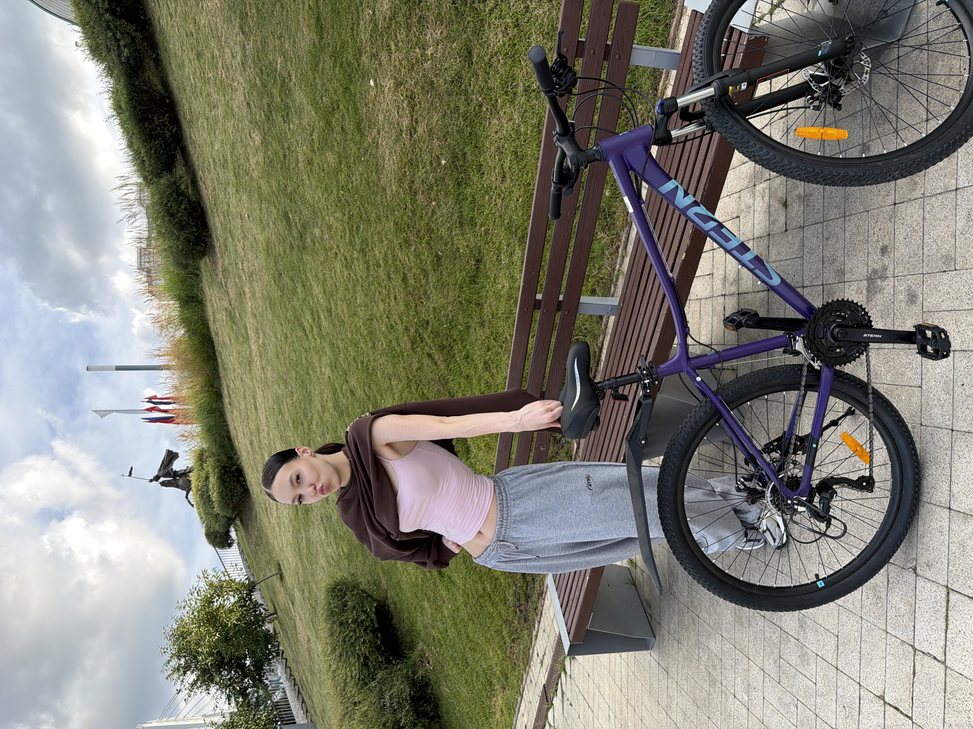 The image shows a young woman with dark hair pulled back in a ponytail, wearing a light pink tank top, grey sweatpants, and a brown sweater draped over her shoulders. She is making a puckered-lip expression at the camera. To her right stands a purple bicycle with the brand name "STERN" visible on the frame. The woman is leaning on the bicycle. The scene appears to be outdoors, with a paved walkway in the foreground, a wooden bench behind the woman, and a grassy slope with some bushes and trees in the background. In the distance, on the top of the slope, there is a statue of a person on a horse, and several flags on poles. The sky is cloudy. The location is identified as being in the city of Krasnogorsk, Russia, possibly near the "Park of Defense" where such a statue and landscape features might be found.