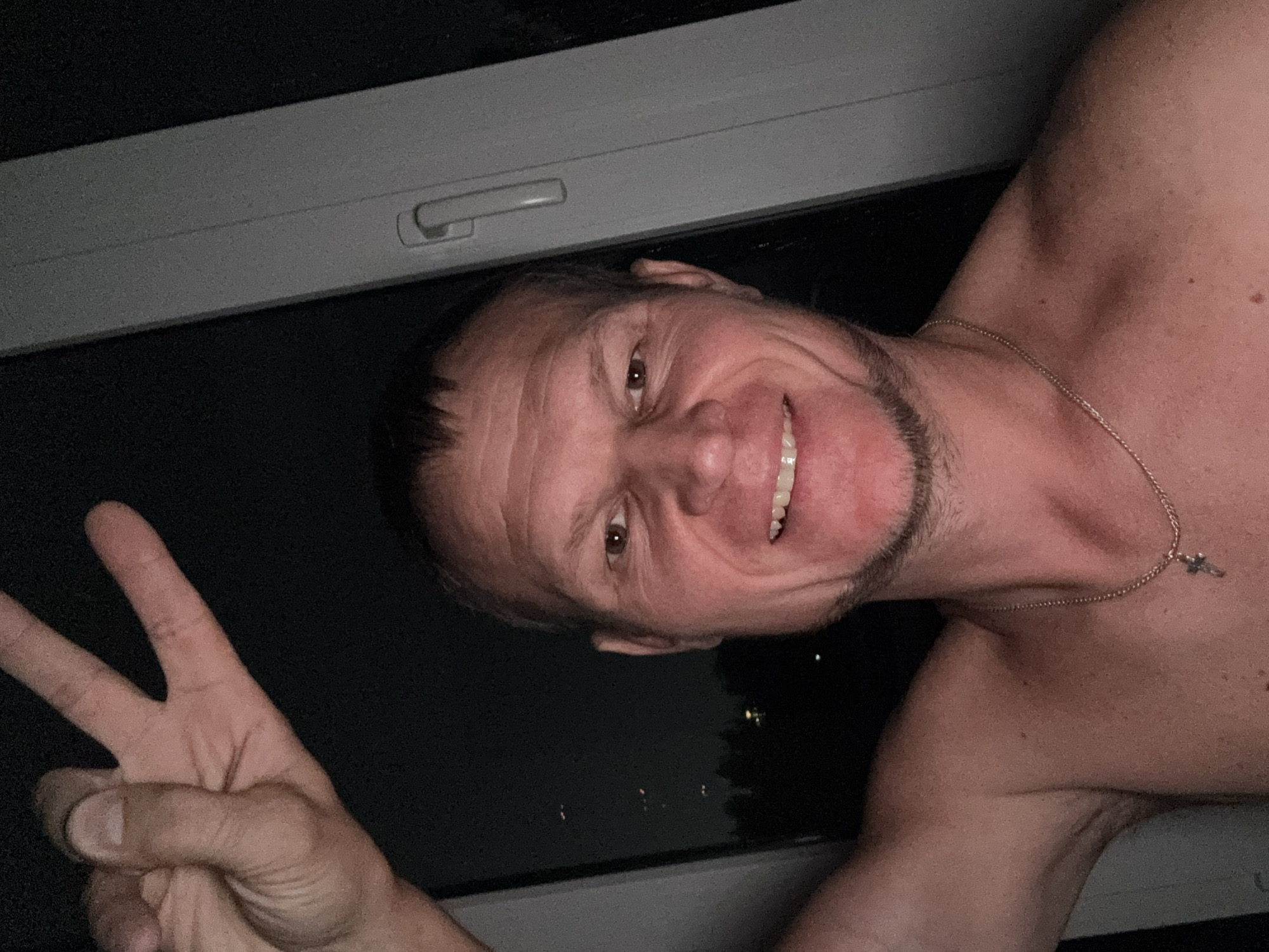 The image shows a shirtless man, appearing to be of Caucasian ethnicity, making a peace sign with his right hand. He is positioned in front of a window frame, which is light gray and has a handle. The background is dark, suggesting it is nighttime, with some faint lights visible in the distance beyond the window, possibly representing city lights or distant structures. The man has short, dark hair, a light beard, and is smiling. He is wearing a thin, silver chain with a cross pendant around his neck. The overall setting appears to be indoors, near a window, with the man taking a selfie. There is no discernible information within the image that can pinpoint its location to Oskemen, Kazakhstan.