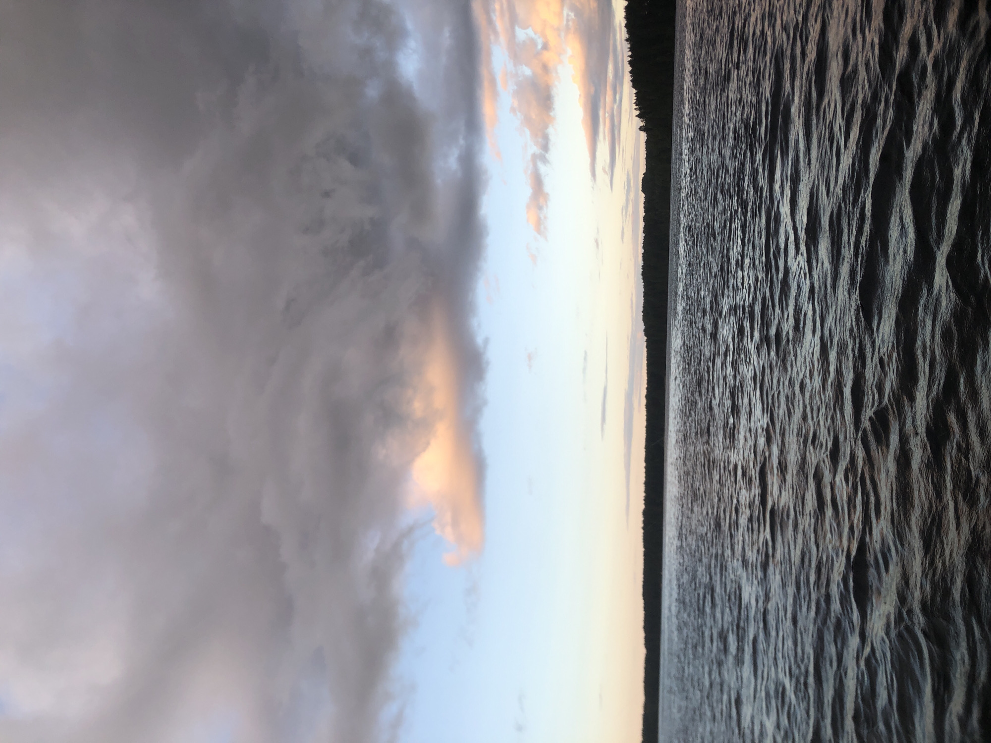 The image depicts a body of water with ripples, likely a lake or river, under a sky filled with dramatic clouds. The clouds are a mix of dark grays and lighter whites, with hints of pink and orange suggesting the soft light of either sunrise or sunset. A dark line of trees forms a silhouette along the horizon on the left side of the frame, separating the sky from the water. The overall scene conveys a sense of natural beauty and tranquility. The time of day is likely dawn or dusk due to the lighting and cloud colors. The location is given as Svirstroy, Russia. There is no visible text in the image.