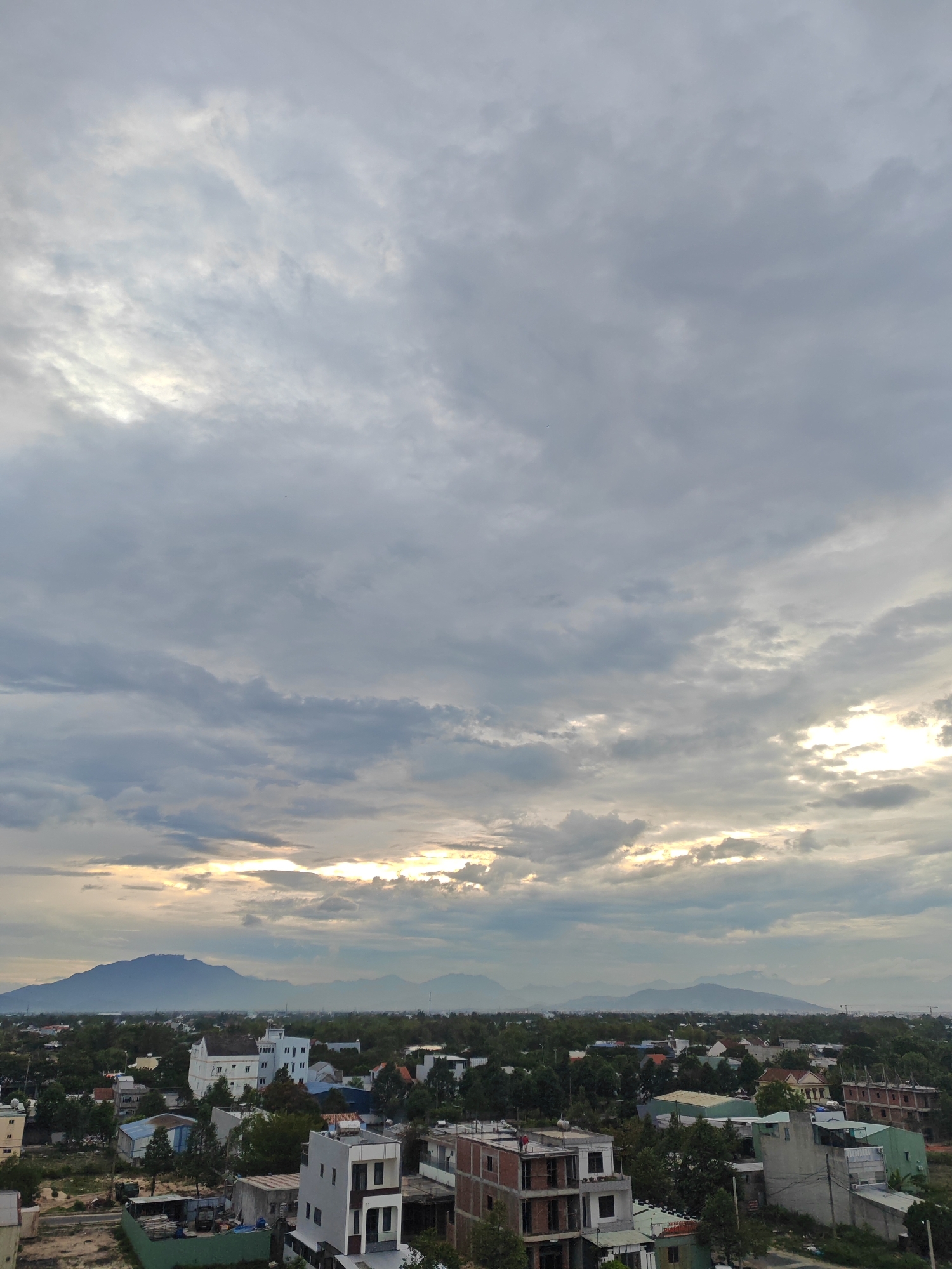 This is an aerial, medium-distance shot of a city during what appears to be sunset or sunrise. The sky is overcast with a mix of dark and light clouds, with patches of sunlight breaking through near the horizon. The light appears to be coming from the west. In the foreground, there are numerous buildings, some under construction, with varying architectural styles. There are also many trees, indicating a significant amount of greenery within the urban landscape. In the background, a range of mountains forms a silhouette against the sky. The scene likely depicts a coastal city in Vietnam, possibly Đà Nẵng, given the presence of mountains in proximity to the sea and the characteristic architecture of residential and commercial buildings. There are no discernible people or active interactions visible in the image. The overall mood is serene and perhaps a bit dramatic due to the cloudy sky.