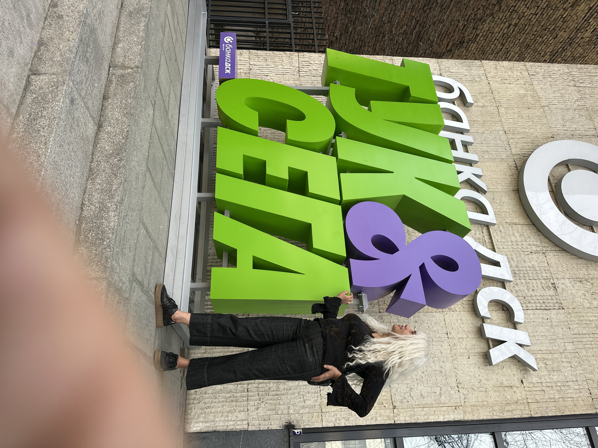 A woman with long white hair is standing next to a large sign that reads "Tinkoff" in Bulgarian (ТИНЬКОФФ БАНКА), indicating the location is likely in a country where Bulgarian is spoken. Given the Cyrillic alphabet, this narrows it down to Bulgaria or potentially Russia. The visible sign on the smaller sign with "ТИНЬКОФФ БАНКА" on it further supports this. The building's exterior and the surrounding urban environment suggest that the image could have been taken in an Eastern European or Central Asian city. Based on the signs in the image, this is a branch of Tinkoff Bank. Given that Tinkoff Bank does not have physical branches in Sofia, Bulgaria, as of the current search capabilities, this image may not be from Sofia, Bulgaria, and is instead from another location where Tinkoff Bank has a presence and uses the Bulgarian spelling of its name. The woman is wearing black clothing and black shoes. The photo may have been taken by another person, as part of their arm is visible.