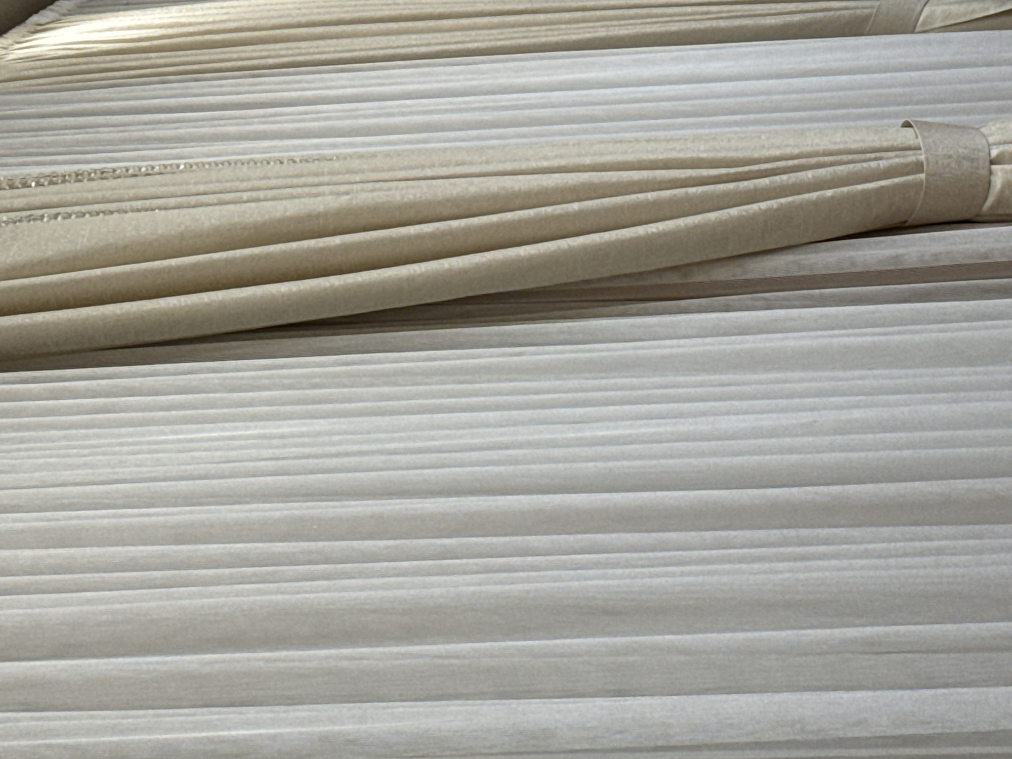 A close-up shot of a window treatment, focusing on its texture and folds. The window treatment consists of two layers of fabric: a sheer, white inner curtain and a thicker, cream-colored outer curtain. The inner curtain is pleated, creating vertical lines that extend from top to bottom. The outer curtain is draped and bunched, with a textured pattern visible on its surface. A decorative beaded trim hangs from the top of the outer curtain, adding a touch of sparkle. The bottom of the outer curtain is secured by a fabric band, also with a subtle pattern. The lighting suggests an indoor setting, likely a room where natural light is being filtered through the window. The scene is static, with no people or actions depicted. The overall impression is one of elegance and soft illumination.