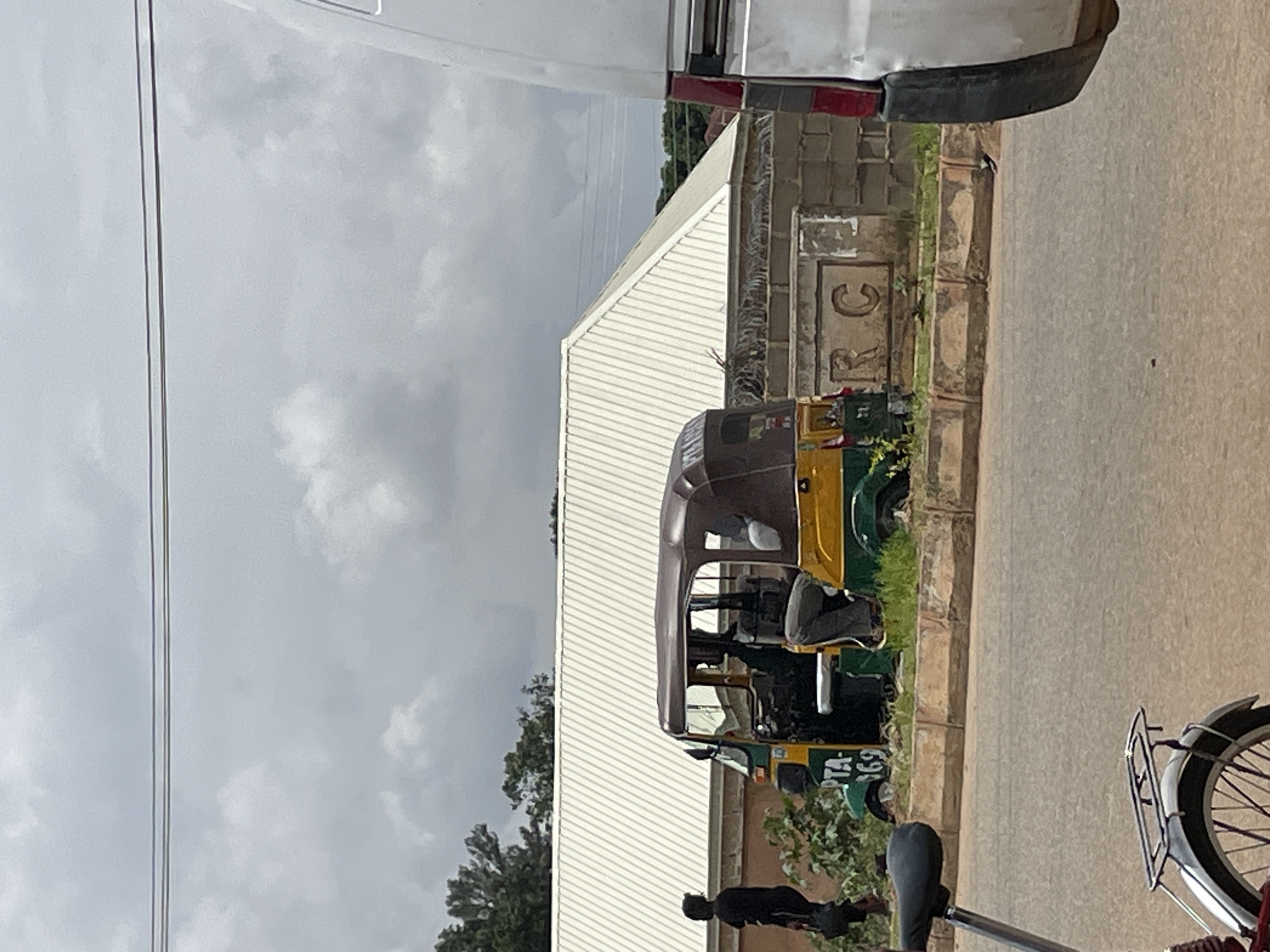 A three-wheeled auto rickshaw, primarily yellow and green with a brown roof, is parked on the side of a road in Bukuru, Nigeria. The auto rickshaw has two passengers visible inside. To the left of the auto rickshaw, a person wearing a black shirt and pants walks away from the vehicle. In the background, a building with a white corrugated metal roof is visible. A portion of a silver vehicle is on the right side of the frame. A bicycle is partially visible in the foreground on the left. The sky is overcast with clouds.