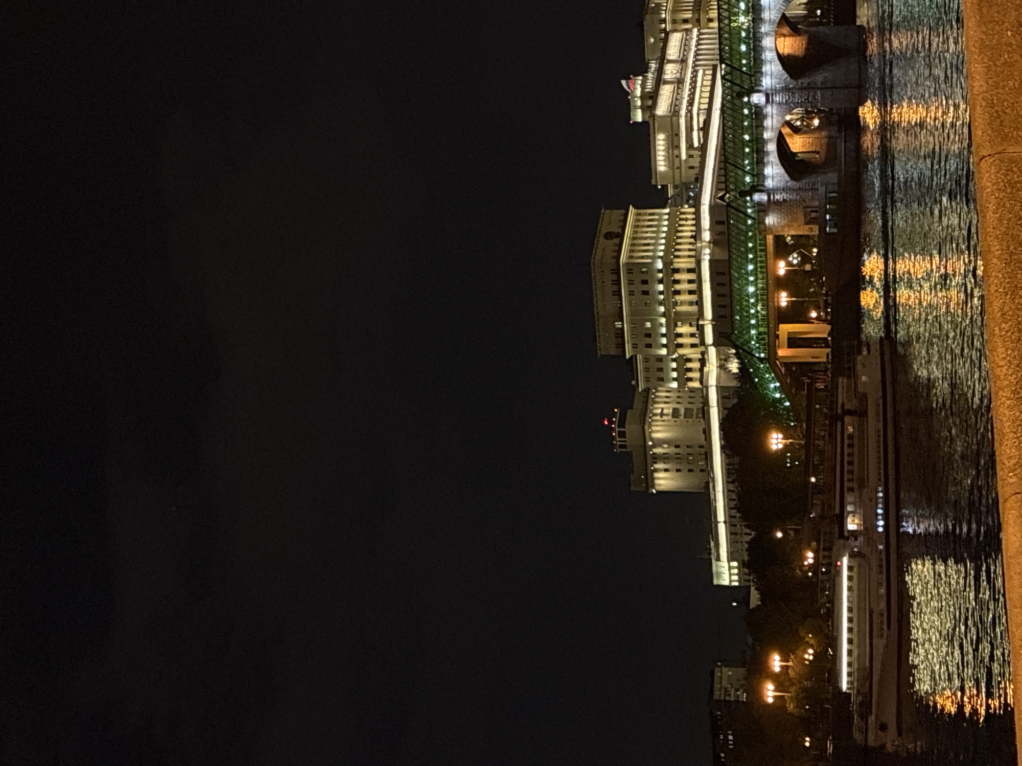 At night, the image depicts a cityscape with illuminated buildings in Moscow, Russia. The structures are adjacent to a body of water, likely the Moskva River, reflecting the lights. A bridge is visible, also illuminated. A boat is also visible along the river. The overall ambiance suggests a serene, urban night scene.