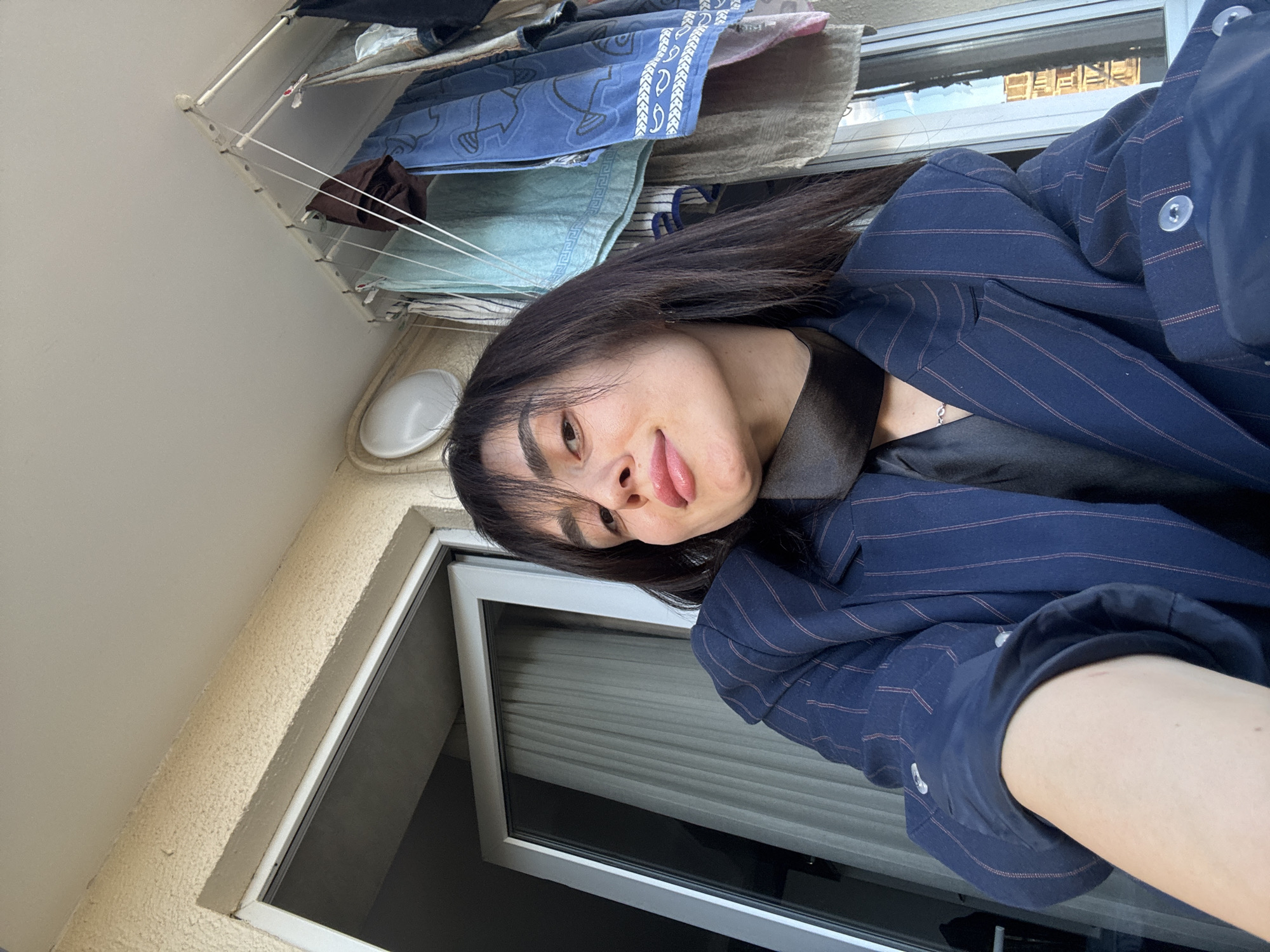 The image is a selfie of a person with long dark hair, wearing a navy blue pinstripe blazer over a black top. The person is facing the camera at an angle, with their tongue slightly visible. The background shows a window with closed blinds, a beige wall, and a clothes drying rack with various items of clothing hanging on it. Given the architectural style and the presence of a clothes drying rack on what appears to be a balcony, the location is likely an apartment in a residential area. The city of Tbilisi, Georgia is not definitively ascertainable from the provided visual details alone.