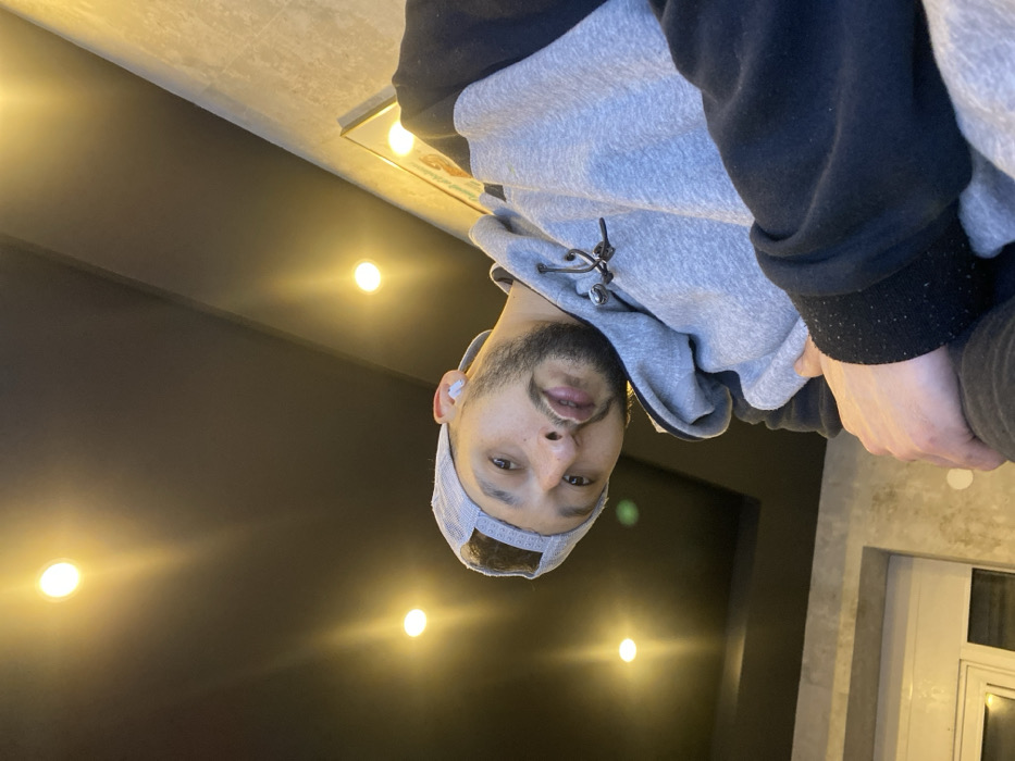 A man in a grey and black hoodie and a grey cap is looking at the camera. He has a beard and an earbud in his left ear. The background is split between a dark wall with overhead lighting and a light grey wall with a white door and window frame. The image is rotated, giving the impression of the man looking down.
