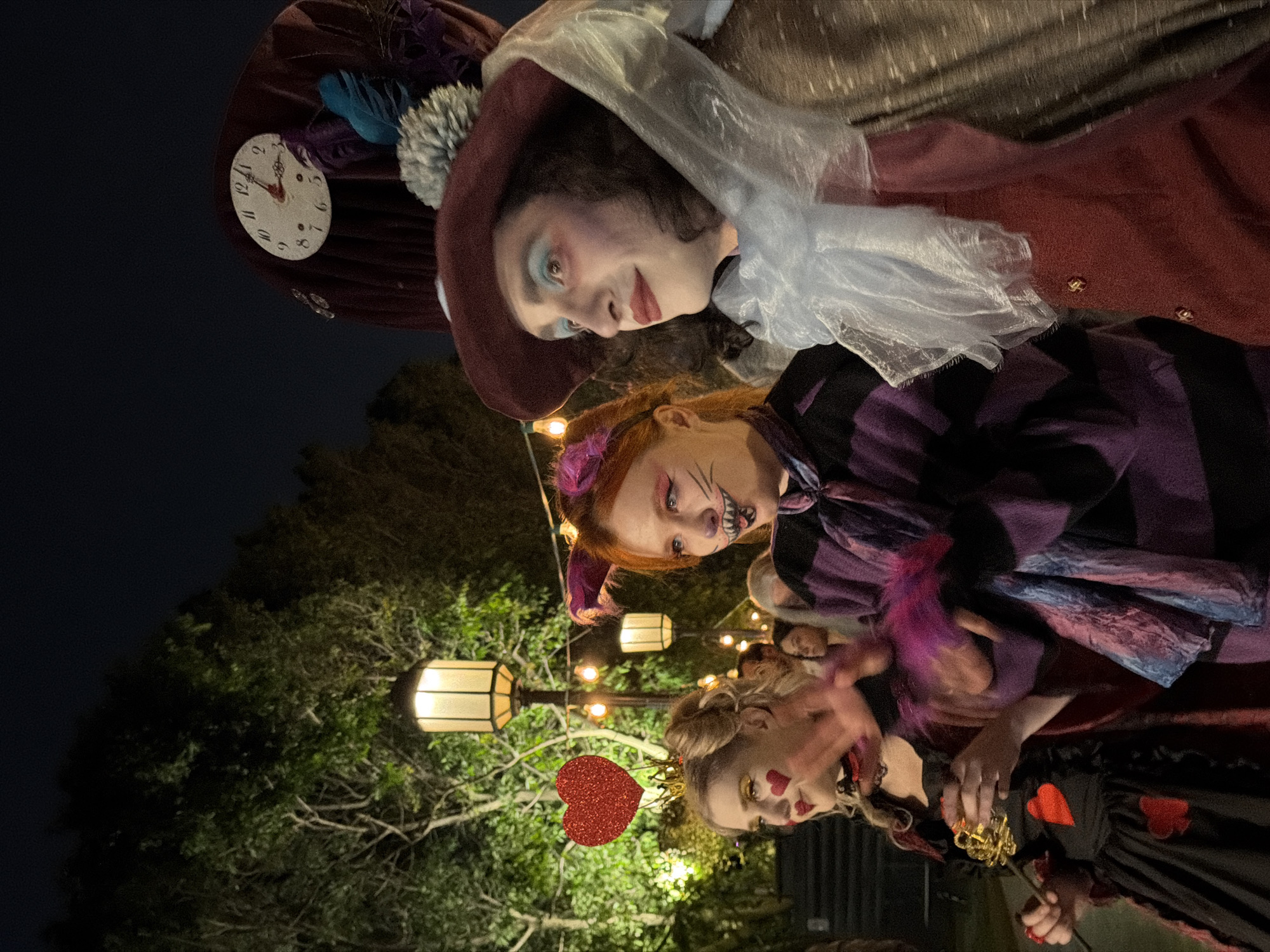 This outdoor night image, captured in Nir Eliyahu, Israel, displays three individuals dressed in elaborate costumes, strongly suggesting an "Alice in Wonderland" theme. They are posing for a photograph in what appears to be a festive, illuminated garden setting.
From left to right (when the image is viewed upright):
The first person is costumed as the Mad Hatter. They wear a reddish-brown top hat adorned with a white clock face, showing hands pointing to approximately 7:00. The hat also features blue and purple feathers and a fluffy grey pom-pom. This individual has pale skin, light blue eyeshadow, and reddish lipstick, and is wearing a reddish-brown jacket with a sheer white ruff.
The middle person is dressed as the Cheshire Cat. They have reddish hair with purple ear-like accessories and prominent cat-like facial makeup, including drawn whiskers and a wide, toothy grin. They are wearing a purple and black striped garment.
The third person on the right is costumed as the Queen of Hearts. They have blonde hair styled up, topped with a small gold crown and a red glittery heart on a headband. Their face is decorated with small red hearts on the cheeks and bold red lipstick. They wear a black dress embellished with red heart designs and are holding a decorative golden key or scepter.
The background is dark, indicating night, with warm string lights and rectangular lantern-style lights hanging amongst lush green trees and foliage, illuminating the scene. The overall mood is whimsical and celebratory, likely from a themed event or party.