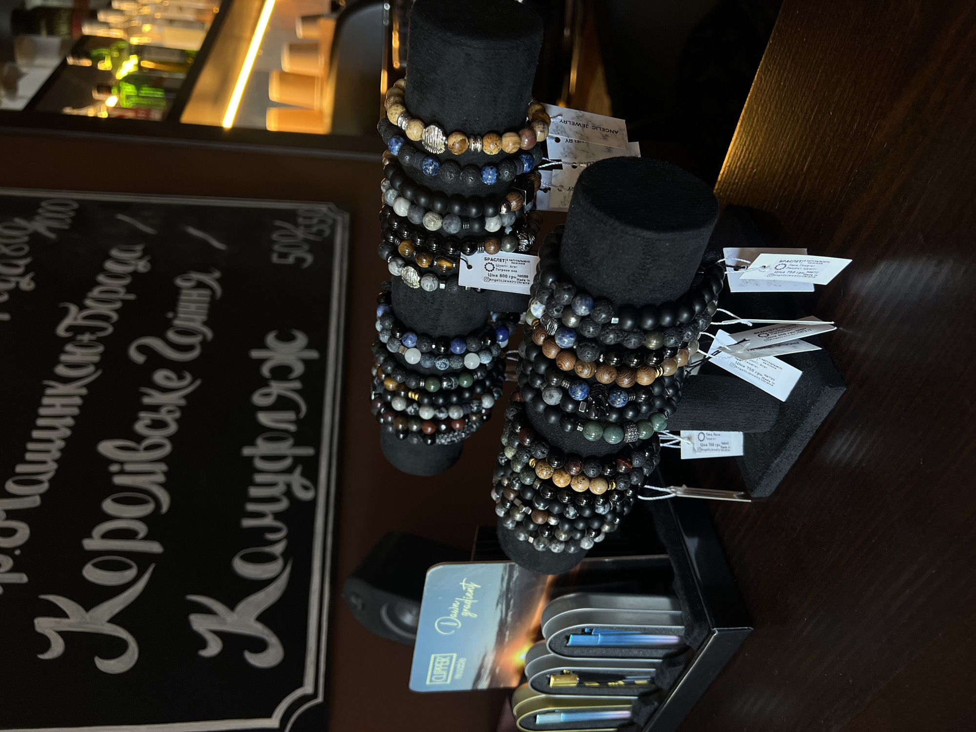 The image depicts a display of beaded bracelets on black stands, arranged on a dark wooden surface. Behind the bracelets, a portion of a black chalkboard with white lettering is visible, featuring handwritten text that appears to be a menu or price list. To the left of the bracelets, a black case holds several colorful lighters and a card with the "CLIPPER" logo and the text "Dawn gradient." In the background, shelves with bottles, cups, and lighting are present, suggesting a bar or retail environment. The overall scene is indoors and appears to be well-lit. The chalkboard's text, which includes Cyrillic script, suggests a location in Ukraine. Based on online search results and local knowledge, this establishment is likely a cafe or bar in Ternopil, Ukraine, potentially named "The Beard Bar," given the mention of "Борода" (Beard) on the chalkboard.