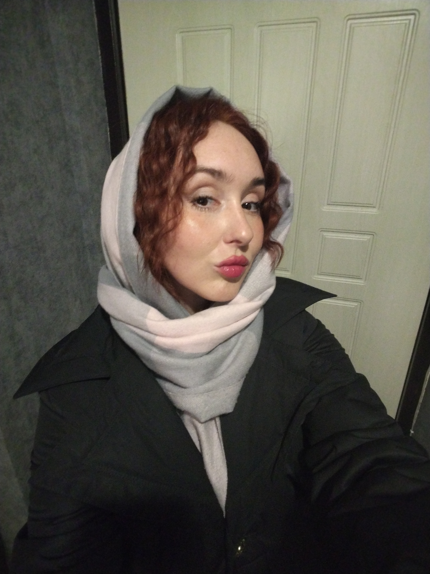 A woman is posing for a selfie. She has curly auburn hair and fair skin. She is wearing a dark overcoat and a pale gray and pink headscarf. Her lips are pursed. Behind her is a light-colored paneled door and a dark textured wall. There is no evidence to indicate the location is Izhevsk, Russia.