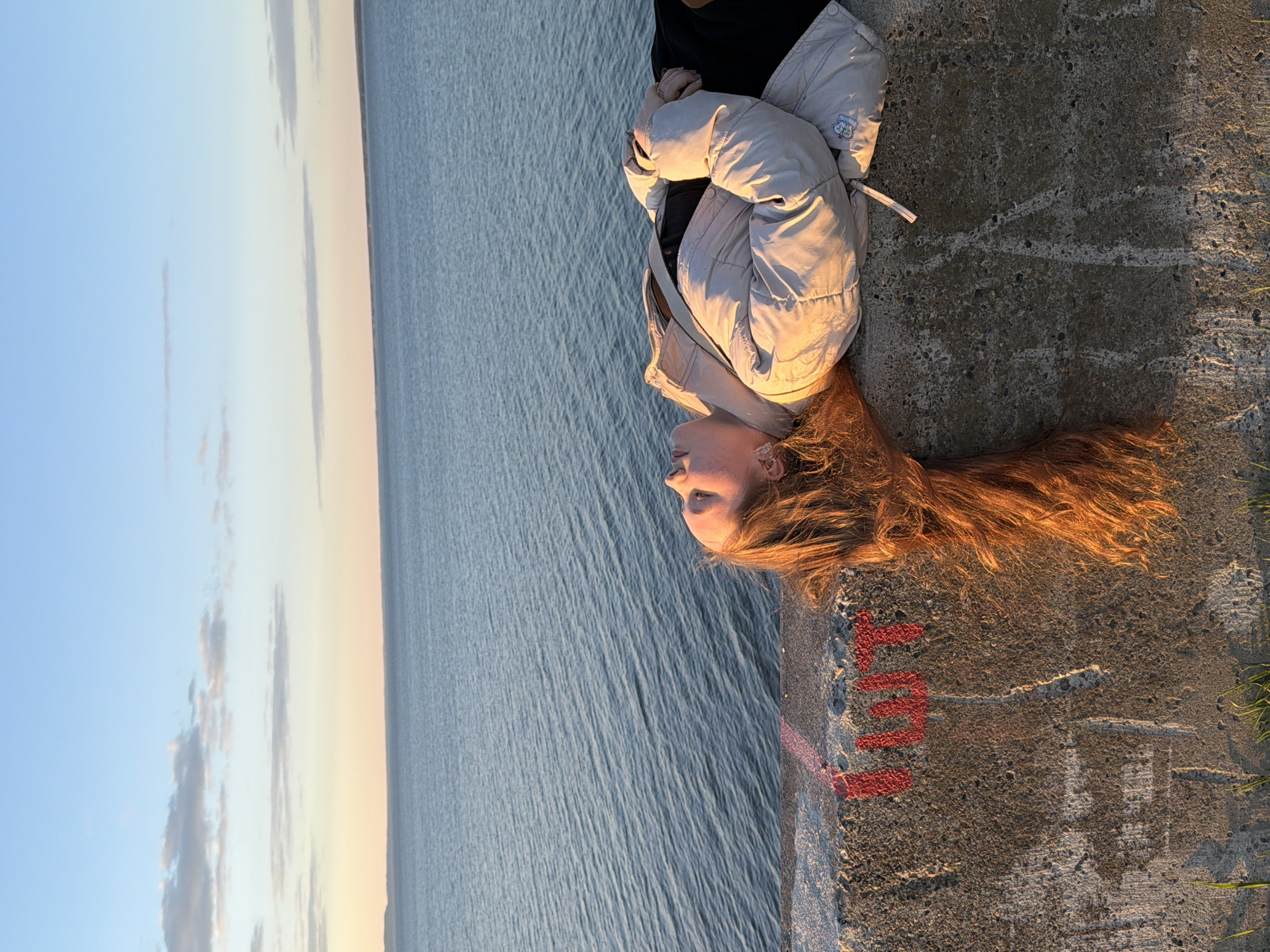 A young woman with red hair lies on a concrete structure, possibly a pier or seawall, overlooking a large body of water. The woman wears a light-colored jacket and dark clothing underneath. The water extends to the horizon, meeting a blue and light-colored sky with some clouds. On the concrete structure near the woman's head, there are red markings resembling letters. Grass is visible along the edge of the concrete. The location appears to be the city of Chaikovsky, Russia based on the waterside construction.