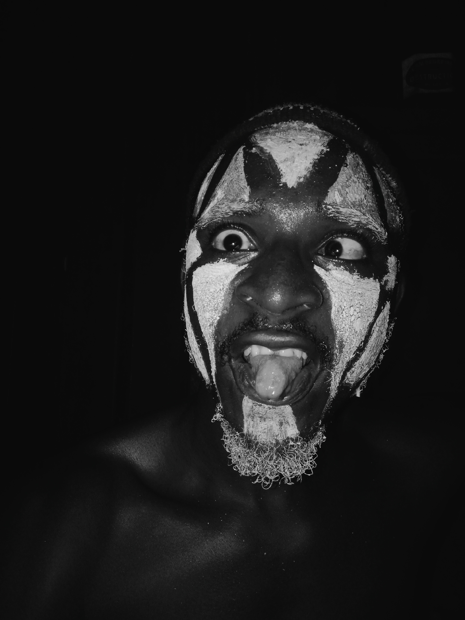 This close-up, black and white portrait features an adult male with dark skin, illuminated against a completely black background. His face is adorned with extensive white paint applied in striking geometric patterns. The white markings form an 'X' shape on his forehead, extend around his wide-open eyes, across his cheeks, and heavily cover his chin and short, textured beard.
The subject displays a highly animated and intense facial expression. His eyes are wide and noticeably crossed, looking distinctly to his right. His mouth is open, revealing his tongue fully extended outwards. This combination of wide, crossed eyes and an open mouth with an extended tongue conveys a sense of wildness, intensity, or a playful, exaggerated emotion.
The lighting is dramatic and high-contrast, strongly illuminating the painted areas of his face and parts of his shoulders, while the rest of the image dissolves into deep shadow. This stark contrast emphasizes the textures of the paint and his skin. The image suggests a performance, ritual, or artistic expression.
The setting appears to be dark, possibly indoors or at night, with a focus on the subject. There is no visible text in the media file. The location context provided is Onitsha, Nigeria.