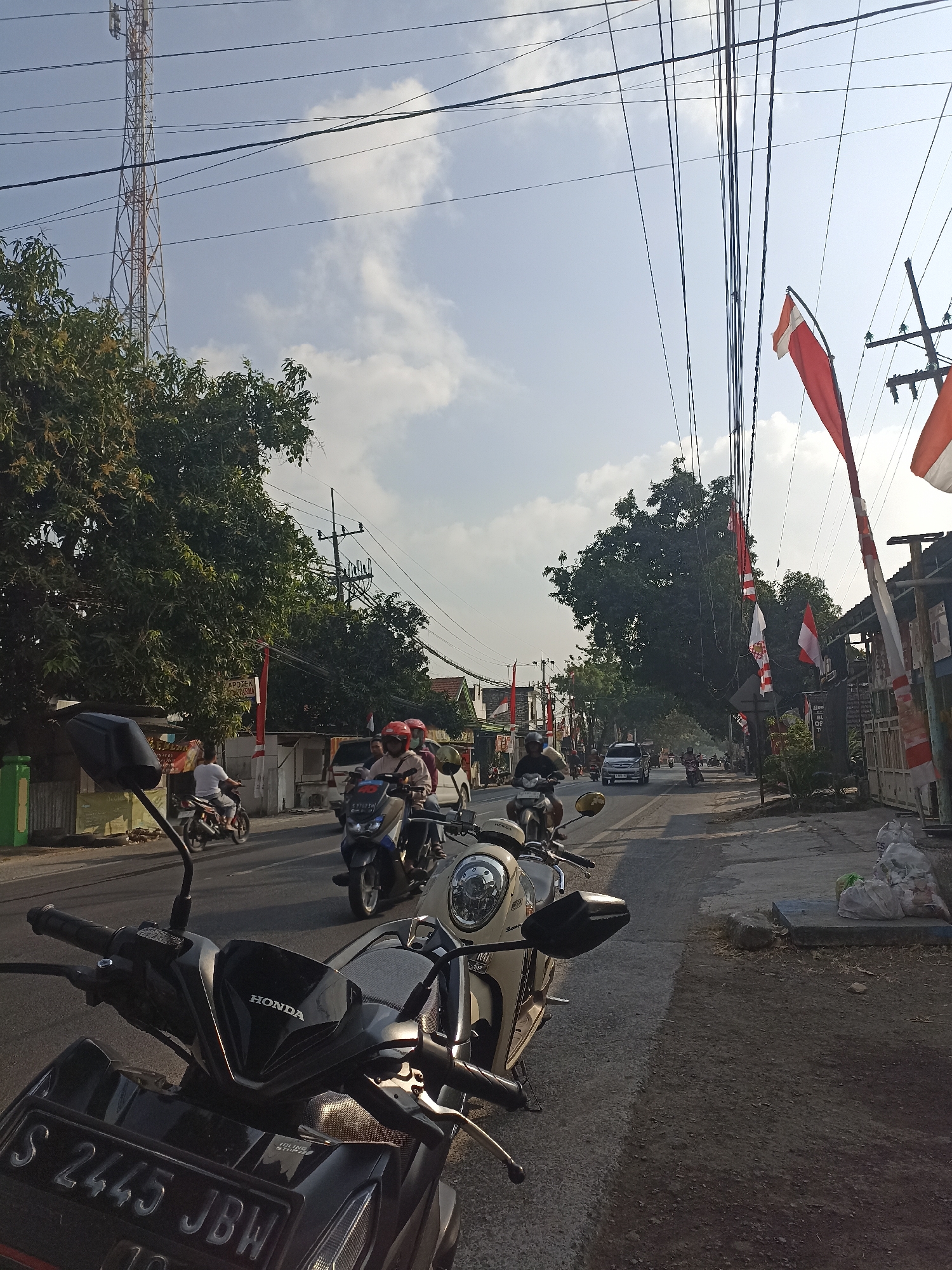 This is a street-level, medium-angle, full-shot photograph taken on a sunny day in Takeranklating, Indonesia. The scene depicts a road with several vehicles and pedestrians. In the foreground, two motorcycles are parked. The one on the left is black with a license plate reading "S 2445 JBW". The one on the right is a cream-colored scooter.

The road itself is paved and appears to be a two-lane street with traffic moving in both directions. Several people are seen riding motorcycles, some wearing helmets. On the left side of the road, there are trees, buildings, and what appears to be a telecommunication tower in the distance. Red and white flags, likely Indonesian flags, are hung along the street, indicating a possible national celebration or event. The right side of the road features more buildings and trees. The sky is blue with scattered clouds. The overall atmosphere is that of a typical day on a moderately busy street in a town or city.