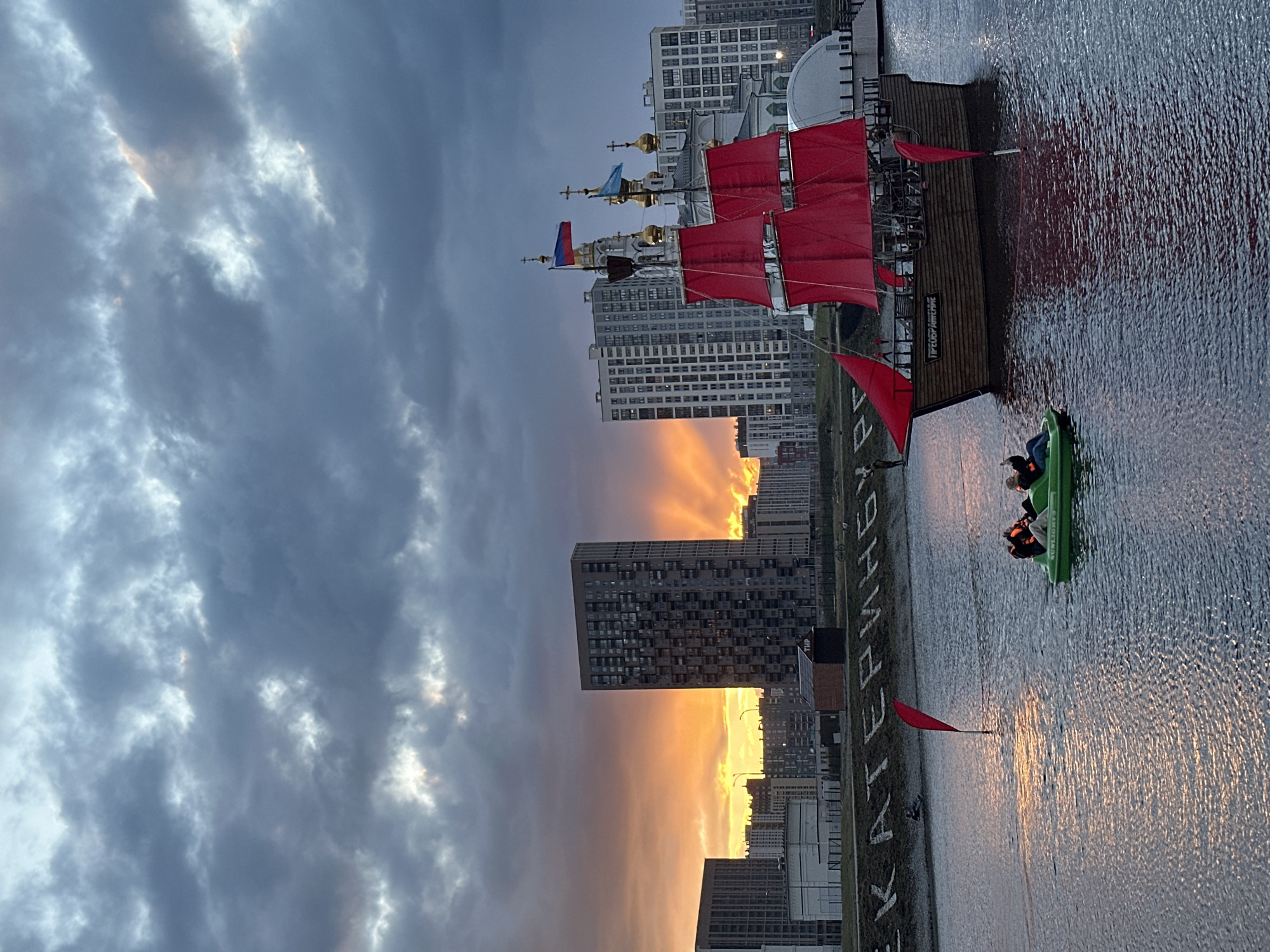 The image captures a twilight cityscape in Yekaterinburg, Russia, oriented vertically.
The scene features a man-made reservoir with a stationary, wooden ship replica with red sails along the right edge. A green paddle boat with two occupants is adjacent to the ship. Ripples are visible on the water's surface, reflecting the sky.
The background reveals a skyline of modern high-rise buildings, partially obscured by a dramatic, layered sky. A vivid orange sunset, peeking through the buildings, contrasts with the dark clouds in the sky. The text "НАБЕРЕЖНАЯ РАБОЧЕЙ МОЛОДЁЖИ" is visible along the embankment, identifying the location as the Embankment of Working Youth.