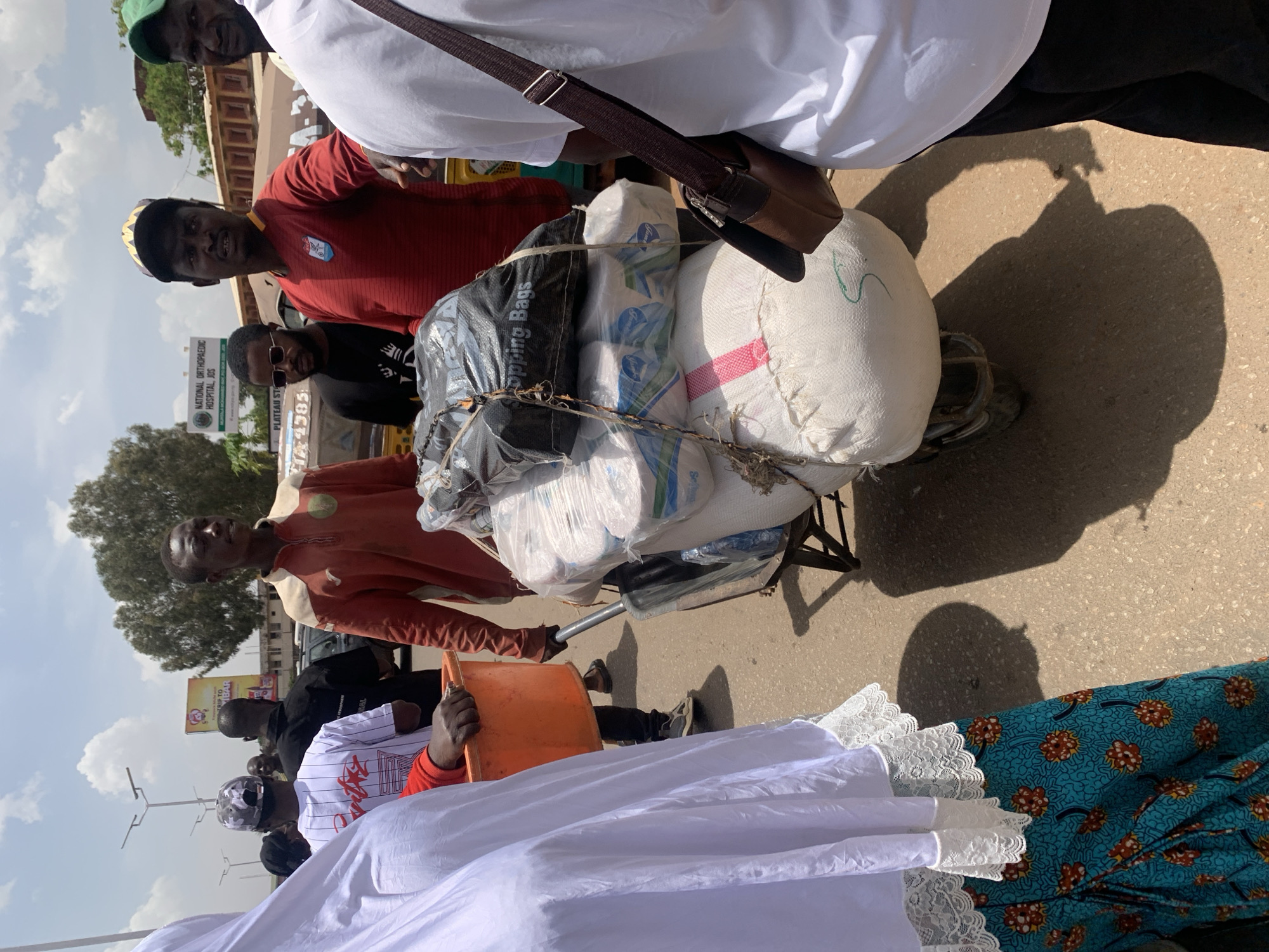 This media file captures a vibrant street scene in Jos, Nigeria, showing several people and a heavily laden cart under bright daylight. The image is oriented vertically (rotated 90 degrees counter-clockwise).
In the center of the frame, a three-wheeled cart or bicycle is piled high with various goods, including large white sacks, a black bag, and bundles of white plastic-wrapped items. One black bag clearly reads "Shopping Bags," and a white bundle shows the brand "Always," likely for hygiene products. One white sack has "Star" partially visible. A man in a light brown or orange long-sleeved shirt stands beside the cart, appearing to push or guide it.
Several individuals are gathered around the cart. In the upper part of the frame, a person in a white shirt and a green cap looks towards the viewer. Below them, a man in a red and yellow striped shirt with a patterned cap also looks towards the camera. Another man with sunglasses and a dark shirt is visible behind him. In the foreground, partially visible, are a person in a white garment with lace trim and a blue patterned fabric, holding an orange bucket.
The setting appears to be an urban street with asphalt pavement. Strong, distinct shadows are cast on the ground, indicating a bright and sunny day, likely midday or early afternoon. In the background, a street sign is visible, reading "NATIONAL ORTHOPAEDIC HOSPITAL, JOS" and "PLATEAU STATE." Further back, a billboard displays the "KFC" logo. Trees and other buildings can also be seen. The scene depicts everyday activity, possibly a bustling market area or goods transportation.