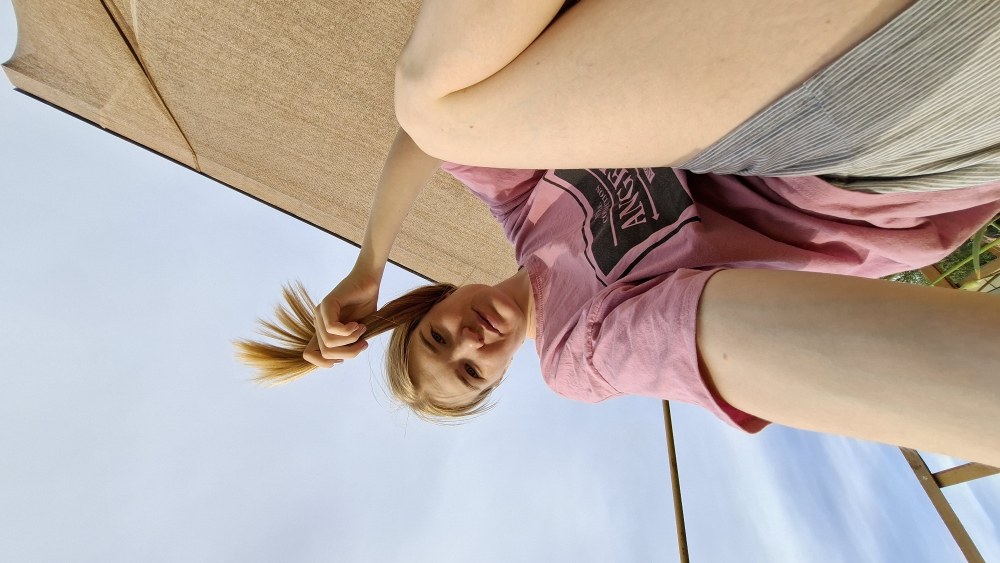 A low-angle shot depicts a woman holding a blonde ponytail under a partially visible beige umbrella. She wears a pink T-shirt with black graphics and grey striped shorts. The background is a blue sky. Elements of an outdoor structure are visible in the lower right corner. The location is speculatively in Большой Букор, Russia.
