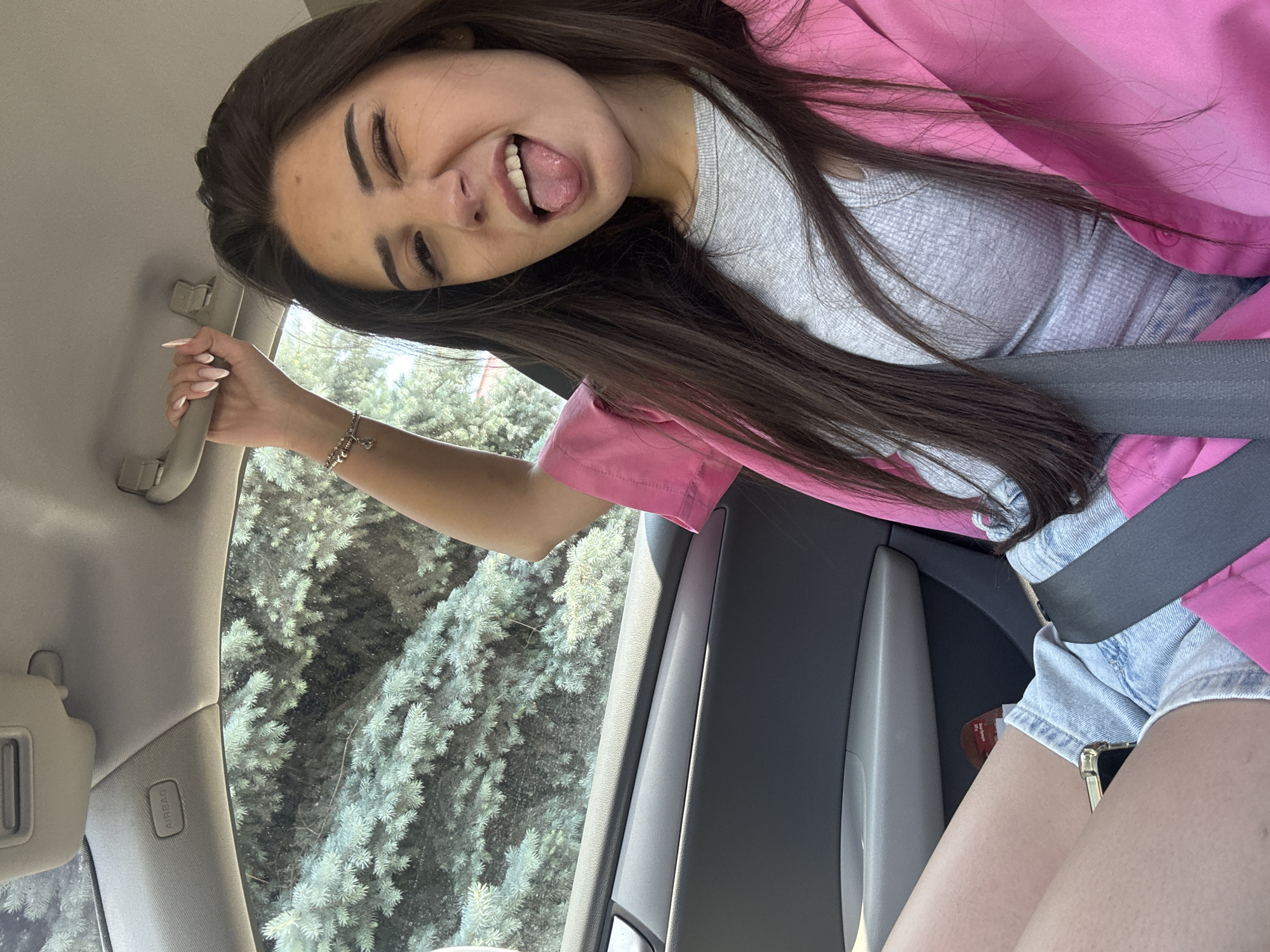 A young woman with long, dark brown hair is captured in a playful selfie inside a car, looking out at a forested area. She is positioned diagonally in the frame, with her head tilted to the right and her body facing slightly forward. Her eyes are squinted in a smile, and her tongue is playfully sticking out. She is wearing a light gray ribbed top underneath a bright pink jacket or shirt, and light blue denim shorts. Her left hand, adorned with long, light pink fingernails and a silver-colored charm bracelet, grasps a gray overhead grab handle inside the car. A dark gray seatbelt is fastened across her chest and lap.
The car's interior is visible, including parts of the light-colored headliner, a gray sun visor, and a panel marked with "AIRBAG" above the window. Outside the car window, a dense cluster of light green evergreen trees, possibly spruce or fir, indicates a natural or planted area. Sunlight illuminates the scene, suggesting it is daytime. A portion of her legs and a smartphone with a gold edge are visible in the lower right corner, with an indistinguishable red tag partially seen near her leg. The overall mood is casual and cheerful, set in Plovdiv, Bulgaria.