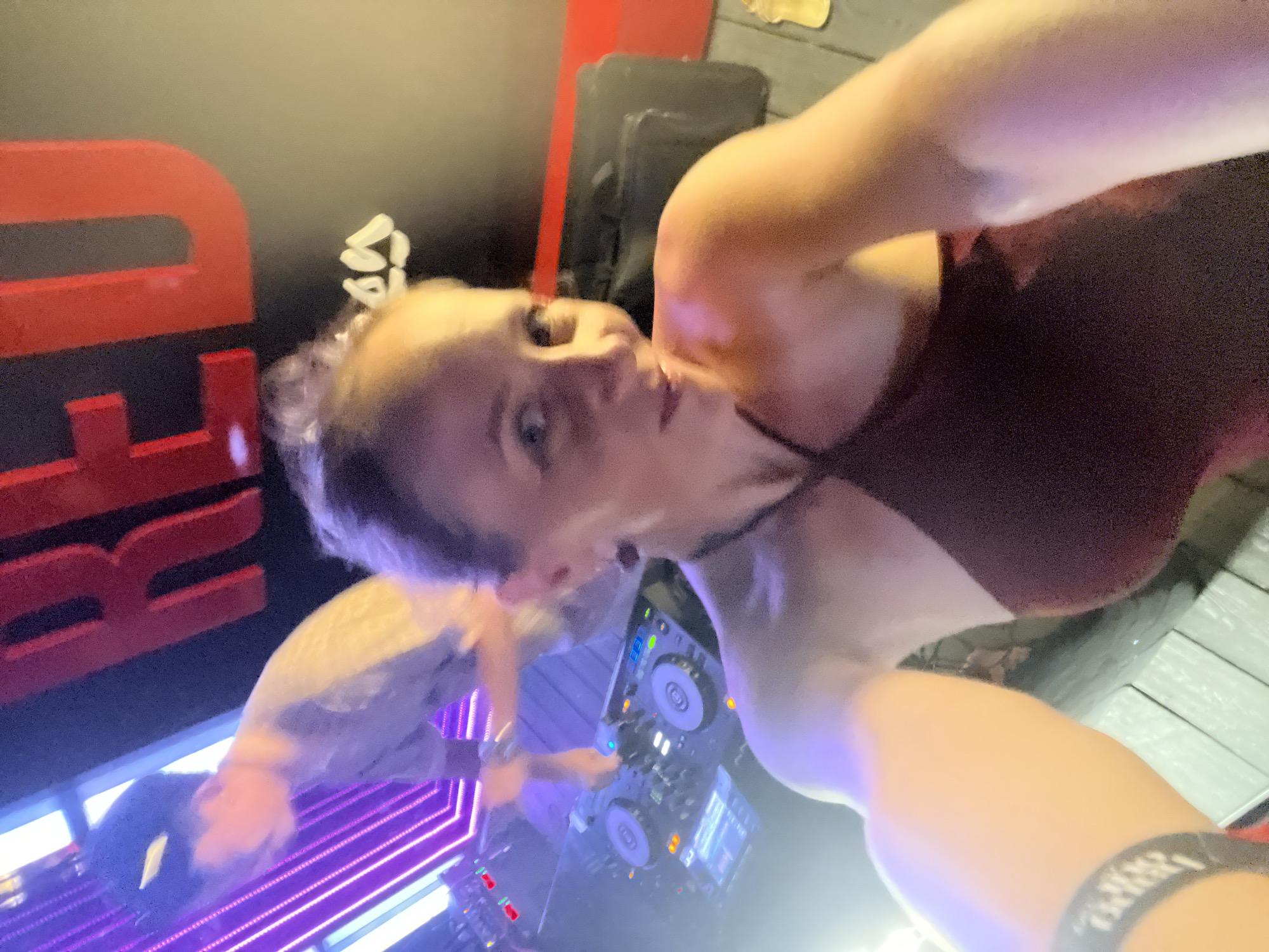 A selfie is taken by a woman in the foreground, wearing a dark red halter top. She is looking at the camera and smiling. Behind her, a DJ set is visible with two turntables and a mixer. A person in a grey shirt is operating the DJ equipment, with their back to the camera. In the background, a large red sign with the word "RED" is illuminated. The scene appears to be in a nightclub or bar, possibly in Da Nang, Vietnam, given the red sign which could be a reference to a popular club name in the city. The lighting is dim and colorful, with purple and white lights contributing to the atmosphere.