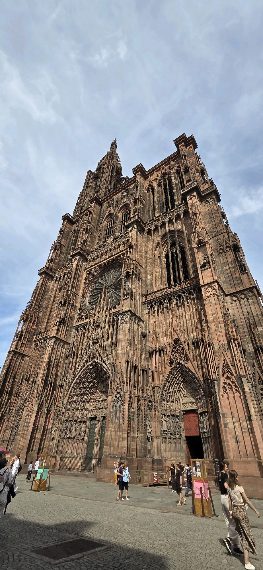 This is a ground-level, wide-angle, vertical photograph of the Cathédrale Notre-Dame de Strasbourg in Strasbourg, France. The sandstone facade of the Gothic cathedral is predominantly red-brown and features intricate carvings, statues, and arched doorways. A large rose window is visible above the central entrance. The sky above is a mix of blue and white clouds. Several people are standing and walking on the cobblestone plaza in front of the cathedral, appearing to be tourists. The lighting suggests it is daytime, possibly late morning or early afternoon, with sunlight casting shadows on the plaza.