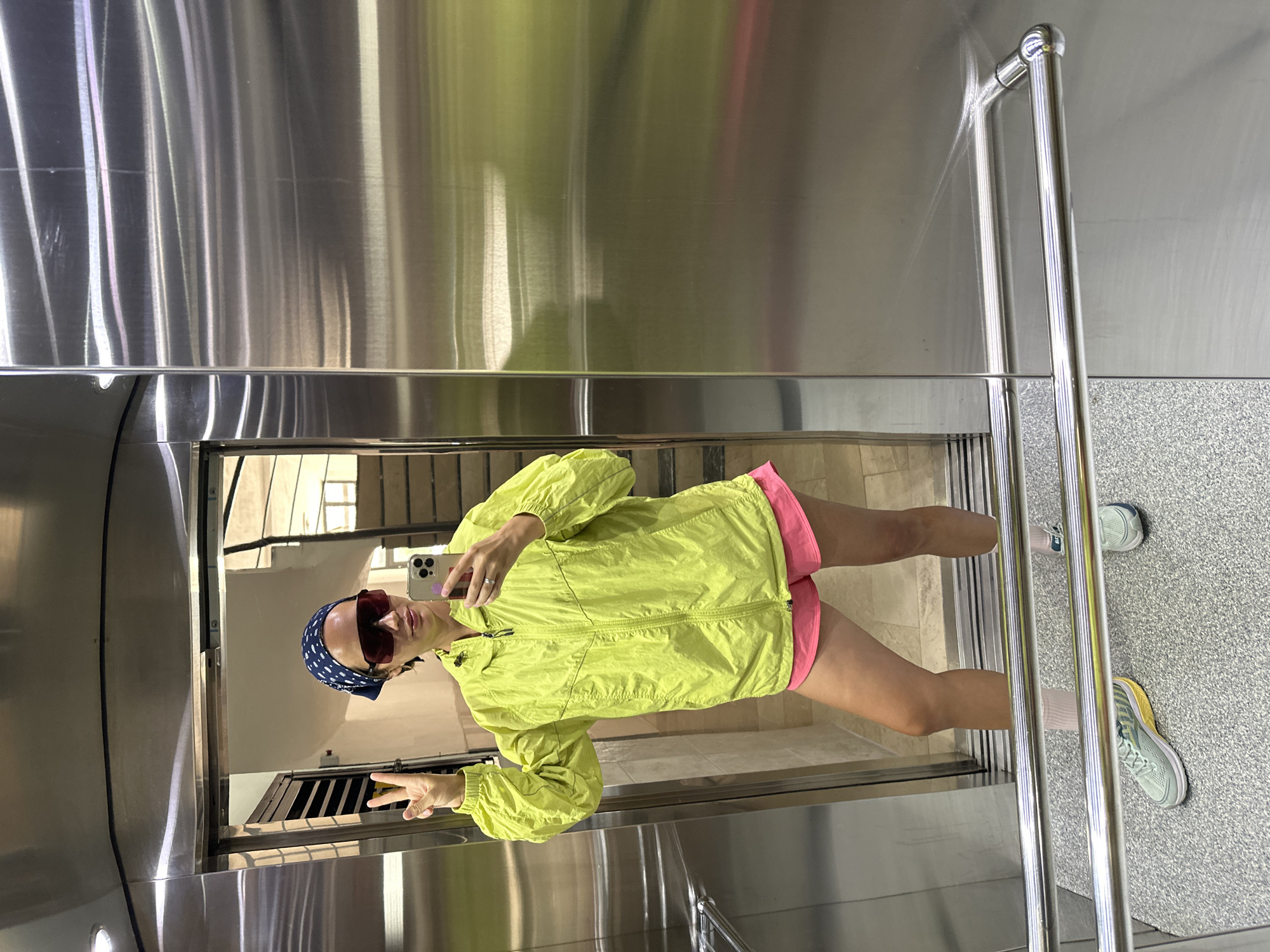 The image shows a person taking a selfie in what appears to be a modern elevator with stainless steel walls. The person is wearing a blue patterned headscarf, oversized sunglasses, a bright yellow zip-up jacket, and pink shorts. They are holding a smartphone in one hand and making a peace sign with the other. The reflection in the elevator shows an interior with stairs and windows. The elevator floor is speckled grey. Near the elevator's metal handrails, two running shoes can be seen. The exact location within Fethiye, Turkey, cannot be determined from the image alone without additional context.