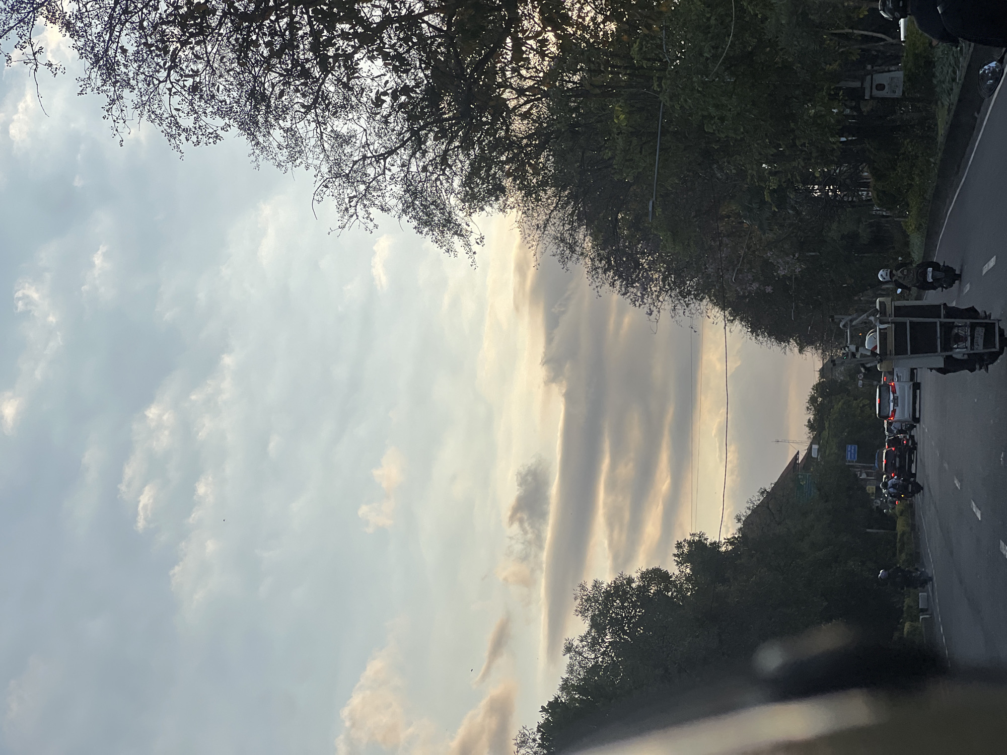 The image captures a street scene in Denpasar, Indonesia, during what appears to be either sunrise or sunset, given the soft, diffused light illuminating the sky and clouds. The perspective is from a vehicle moving on a road lined with dense foliage and trees.
In the foreground, the left side of the frame is partially occupied by the interior of a vehicle, possibly a car, with a dark, reflective surface visible. The road ahead shows moderate traffic, including several cars and numerous motorcycles. One motorcycle in the center of the frame is carrying a large, ladder-like structure on its back, with two individuals wearing helmets. Other motorcycles are also visible with riders, suggesting typical urban traffic in the area.
The road is paved and has lane markings. On the sides of the road, there are sidewalks, vegetation, and trees with dense canopies, some of which are silhouetted against the sky. In the background, buildings with reddish-tiled roofs are partially visible through the trees. The sky is a mix of light blue and pale yellow, with scattered white and gray clouds, some of which are catching the warm light, creating streaks of color. The overall atmosphere is calm and transitional, indicative of either the start or end of the day in a tropical urban setting.