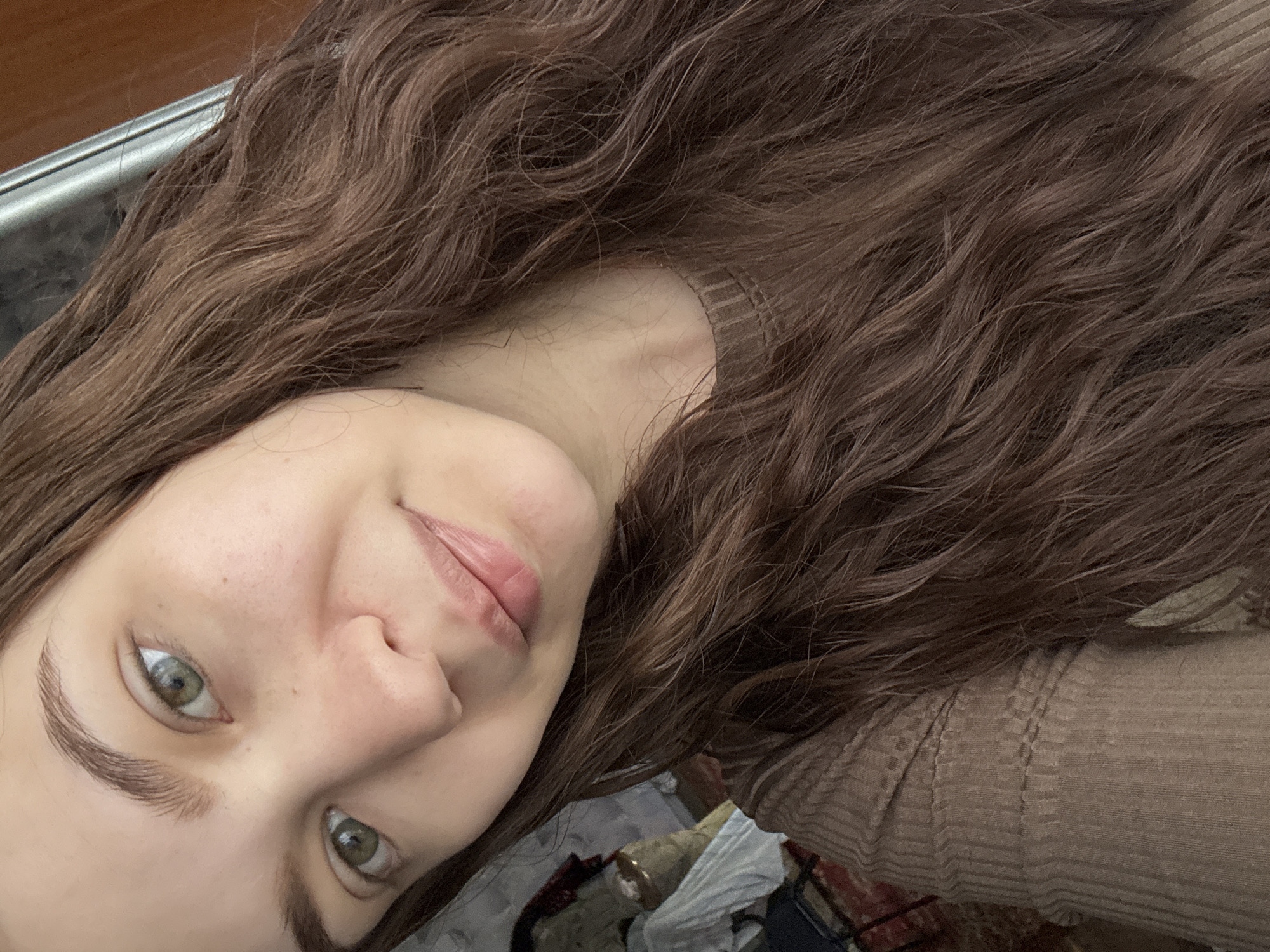 The image features a young woman with long, wavy brown hair. She is wearing a beige sweater. Her skin is fair, and she has light green eyes and defined eyebrows. She appears to be in an indoor setting, with the corner of a metal object visible in the upper left. The background is cluttered with indistinct objects. There is no indication of the location being in the city of Nizhnyaya Gar', Russia.