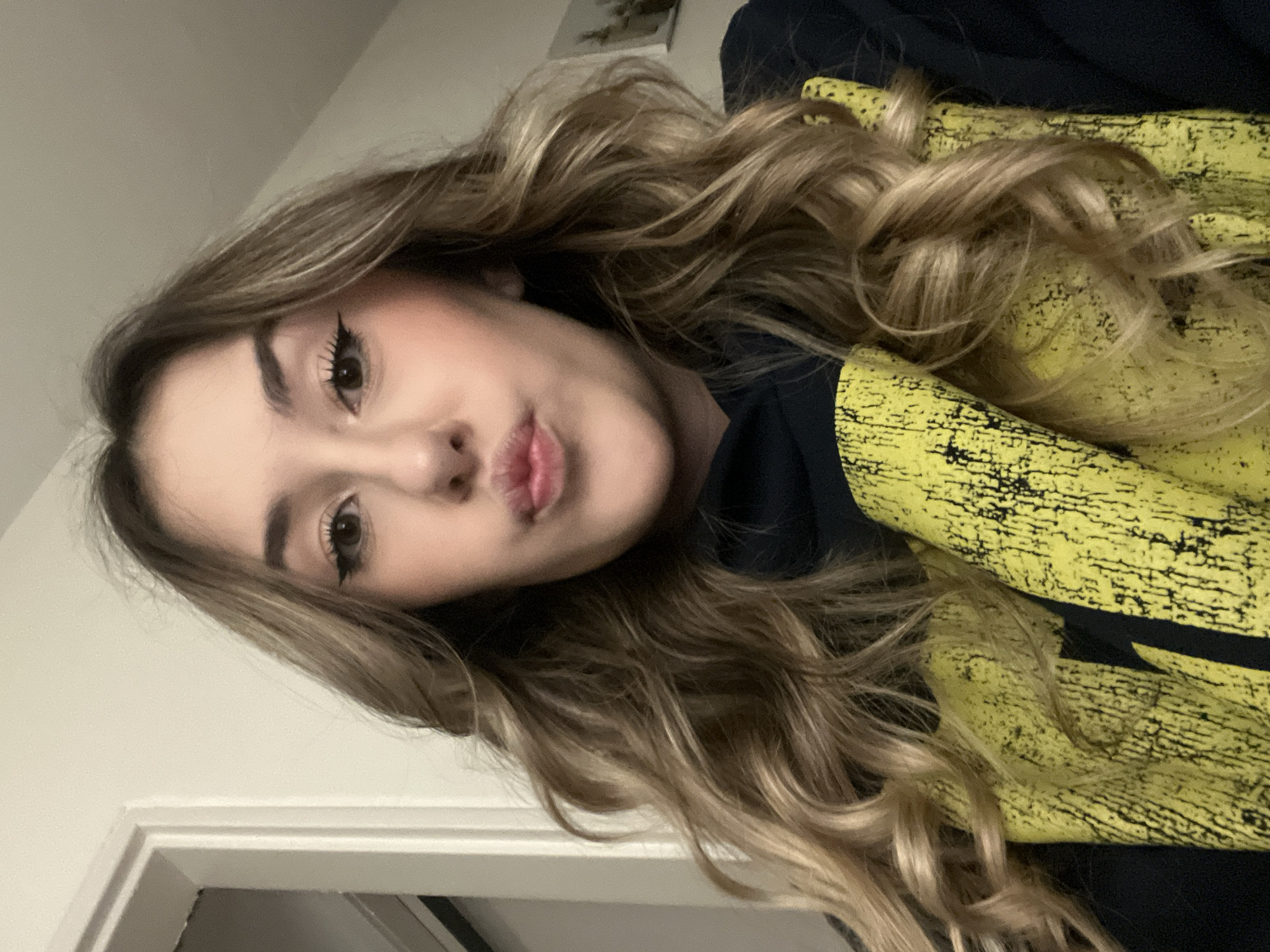 A young woman with long, wavy, light brown hair is posing for a selfie. She has light skin, brown eyes with winged eyeliner, and is making a kissing face. She is wearing a yellow and black hooded sweatshirt. The background appears to be an interior setting with a white wall and a door frame visible. There is not enough information in the image to determine a specific location in Sofia, Bulgaria.