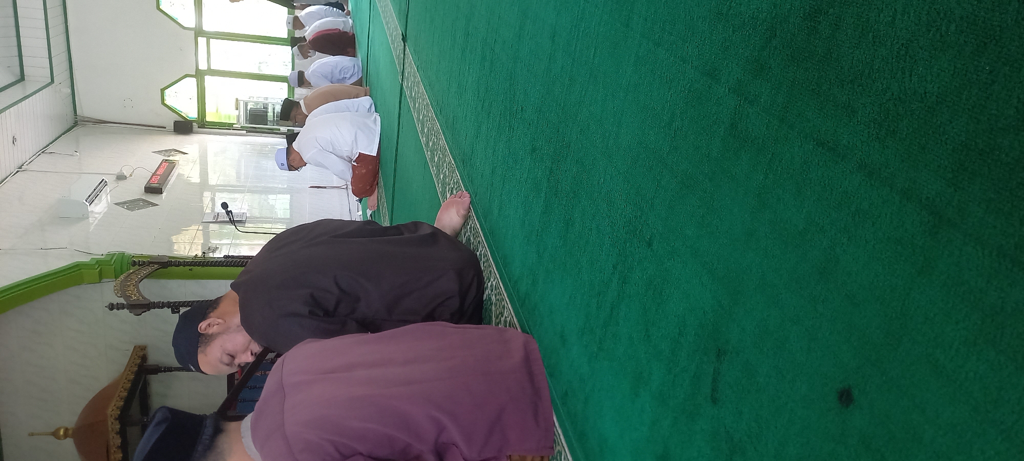 The image depicts a group of men sitting on a green carpet inside a mosque in Loa Lepu, Indonesia. In the foreground, the back of a man wearing a dark robe and a black skullcap is visible. To his left, another man in a purple garment is also seen from behind. Further into the mosque, several men are seated in rows, facing forward. They are dressed in traditional Islamic attire, including white shirts and skullcaps, and some are wearing sarongs. One man in the second row from the front is reading from a book. The environment suggests a prayer or religious gathering. The mosque's interior features white tiled walls, a green-trimmed ceiling, and windows with green frames. A digital clock displaying "12:13" is mounted on the wall, along with an air conditioning unit. A microphone is positioned on a stand in the middle ground.