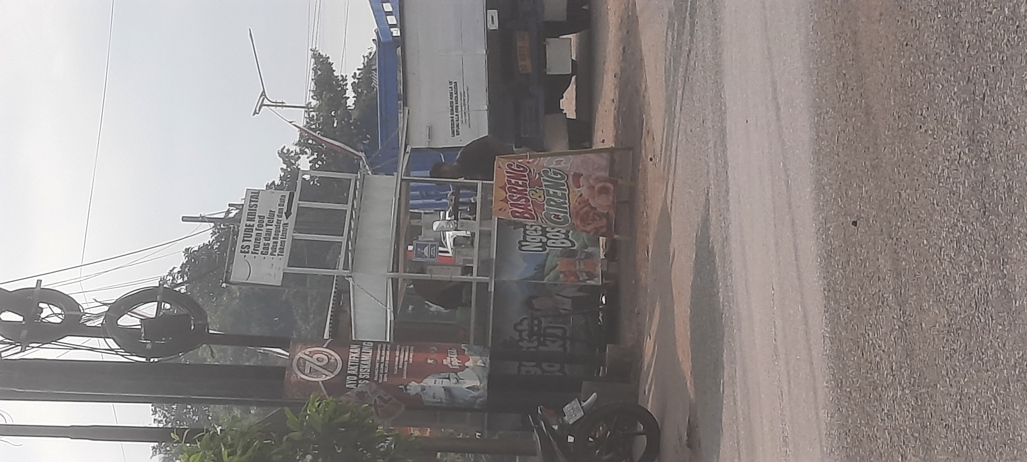 The image depicts a roadside stall in Siraman, Indonesia, with a focus on a small food and drink stand. The stall is set up under a white canopy with various signs. One sign advertises "ES TUBE KRISTAL," "frozen Food," "Gas dan Telur," "Pulsa Regular dan Data," and "Token Listrik." Another sign on the left, featuring the number "76" in a red circle, urges to "AYO AKTIFKAN BOS SISKAMLING." Below this, there is an image of a man in uniform and text that is partially obscured.

In front of the stall, an A-frame sign displays an advertisement for "Nges BASRENG & BOS CIRENG," with images of fried food items. A motorcycle is parked to the left of the stall. Behind the stall, a large blue truck is visible, and a person in a dark shirt stands near it. The environment appears to be a street with a paved road in the foreground and trees in the background under an overcast sky. The overall scene captures a typical street vendor setup in an Indonesian locale.