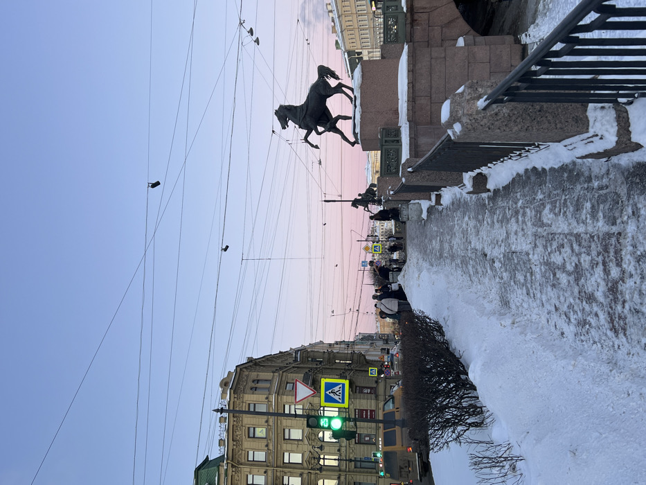 The image displays a winter scene in Saint Petersburg, Russia, centered on the Anichkov Bridge over the Fontanka River. The sky transitions from a pale blue at the zenith to a soft pink and lavender hue near the horizon.
In the foreground, a paved pedestrian walkway, partially cleared of snow and exhibiting icy patches, extends from bottom center towards the middle ground. To the left of the walkway is a mound of undisturbed snow, bordering a section of bare, deciduous shrubbery. To the right, a stone and dark metal railing, also partially covered in snow, delineates the edge of the Anichkov Bridge.
Two of the "Horse Tamers" equestrian sculptures by Peter Clodt von Jürgensburg are visible on the bridge. The nearest statue, a dark-colored horse rearing on its hind legs with a human figure beside it, stands on a light-colored stone pedestal. A second, more distant statue of a similar motif is visible further along the bridge.
On the left side of the frame, a multi-story, ornate building with light-colored masonry and a green roof stands at a street corner. Below this building, a traffic light displays a green signal for vehicles and a white illuminated pedestrian symbol. Associated road signs, including a triangular yield sign and a blue square pedestrian crossing sign, are present. A light-colored van is partially visible on the street below the traffic light, with other vehicles further down the road. Approximately ten indistinct pedestrians are clustered on the sidewalk near the building corner.
Numerous dark utility and tram wires crisscross the upper half of the image against the sky. Two dark, shoe-like objects are suspended from separate overhead wires. Buildings are visible on the far bank of the Fontanka River beyond the bridge.