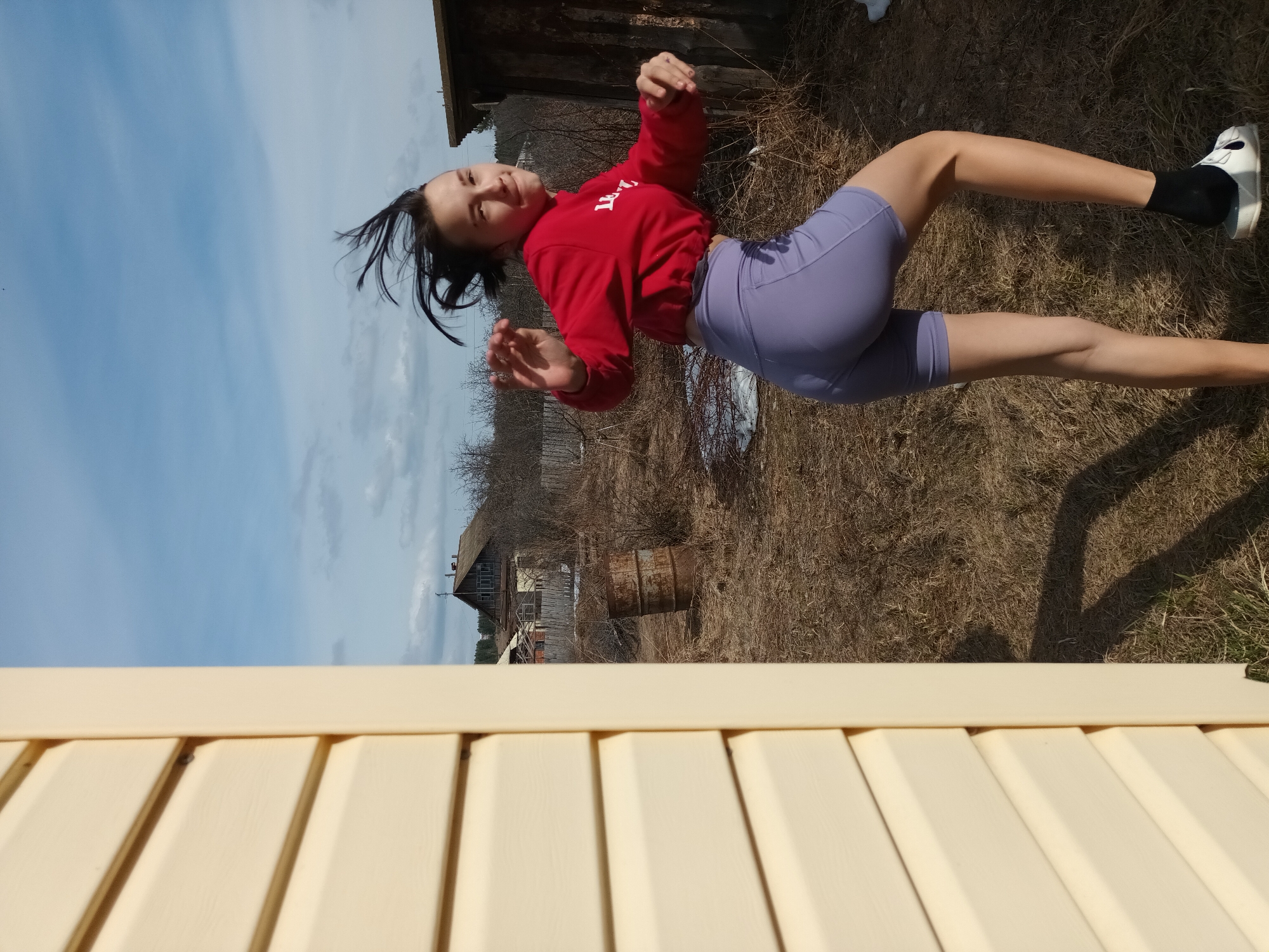 A young woman in a red sweatshirt and purple shorts is suspended horizontally in the air, facing the viewer. Her hair is loose and slightly disheveled, and she is wearing black socks and white shoes. The background consists of dry grass, bare trees, and a wooden building with an old barrel nearby. A clear sky with some clouds is visible in the upper left. A light-colored wooden fence is in the foreground. The location is Ольховка, Russia.
