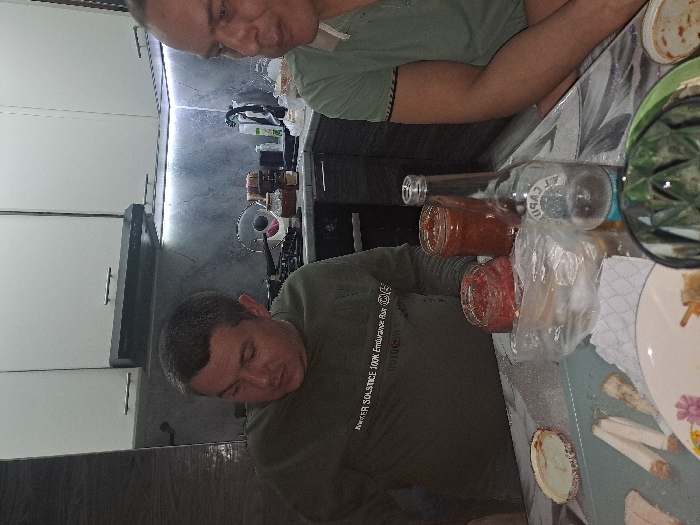 Two men are sitting at a table laden with food and condiments. A bottle of alcohol lies on its side near jars of what appears to be homemade salsa. Plates with food remnants are scattered across the table's surface. The setting is a kitchen, indicated by the presence of cabinets and countertops in the background. The image is slightly tilted.