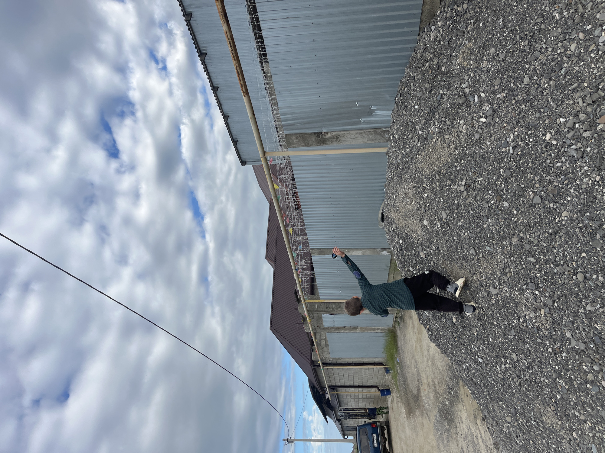 A person stands on a gravel surface beside a building in Kaspiysk, Russia. The building has a corrugated metal facade and a brown roof. The person is wearing a green shirt and black pants and appears to be holding an object in their raised right hand. Above the building, the sky is filled with white and gray clouds. A utility pole and a blue van are also visible in the background.