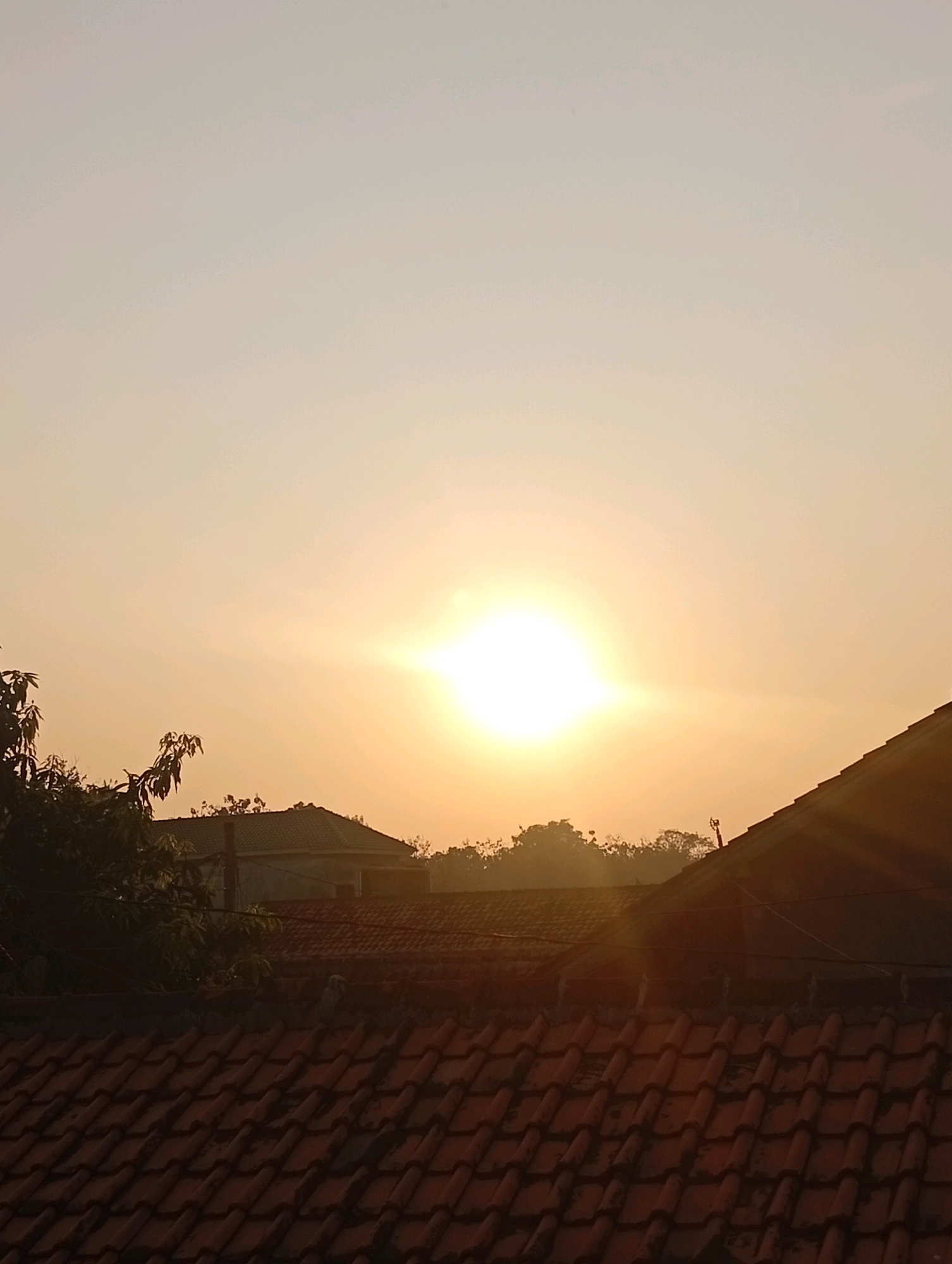 The image shows a sunrise over a residential area in Kebandingan, Indonesia. The sun is a bright, glowing orb in the sky, casting a warm, orange light over the scene. In the foreground, there are several rooftops made of red tiles, some of which appear to be slightly weathered. Behind the rooftops, there are trees and the roofs of houses. The sky is a gradient of pale yellow and orange, indicating the early morning light. The overall impression is one of a peaceful, rural sunrise.