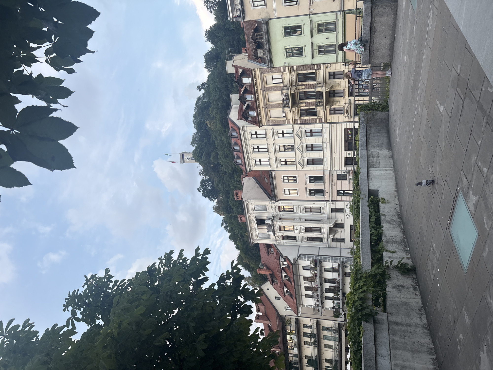 In the city of Unknown, Unknown, a stone walkway borders a canal, with lush green vines growing over a concrete barrier. Beyond the barrier, a row of colorful, multi-story buildings with red tiled roofs line the waterfront. The buildings exhibit diverse architectural styles, some featuring balconies and ornate window details. In the background, a densely forested hill rises, topped by a prominent clock tower flying two flags. On the walkway, two young girls stand near the barrier, looking out at the scene. A single pigeon stands on the pavement in the foreground. The sky above is partly cloudy.