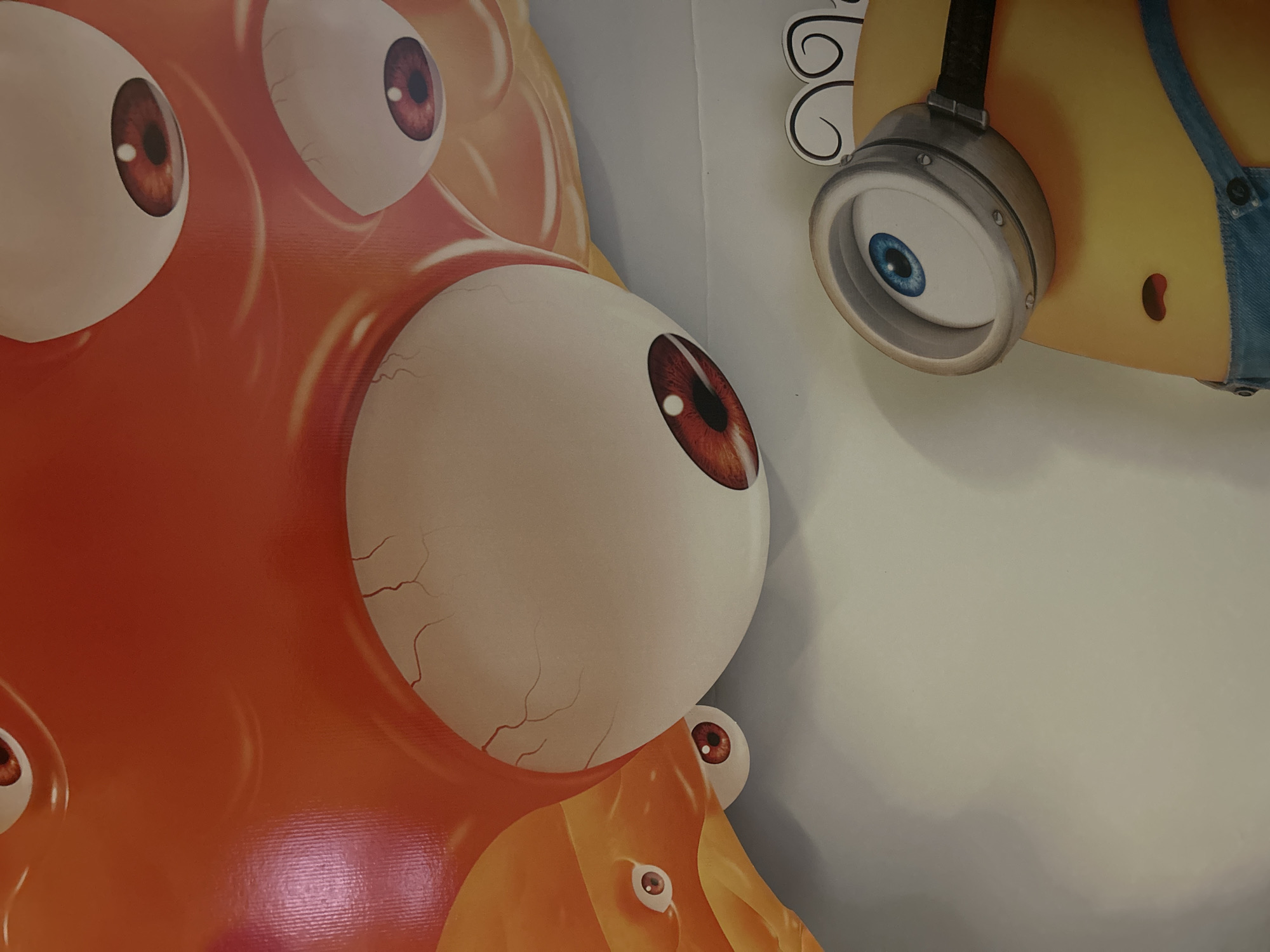 The media file is a close-up photograph of a cartoonish, orange, blob-like creature with multiple large, white eyeballs. The creature's eyes are detailed with reddish-brown irises and black pupils. One of the eyes is particularly large and dominates the foreground of the image. In the background, a yellow character, resembling a Minion from the "Despicable Me" franchise, is partially visible. This character is wearing a silver goggle with a blue iris. The overall scene appears to be a display or decoration, possibly in a public space, given the large scale of the cartoon characters. The lighting suggests an indoor environment. There is no visible text within the frame. The context provided indicates the location as Byala Slatina, Bulgaria, which might imply this is part of a local event or attraction.