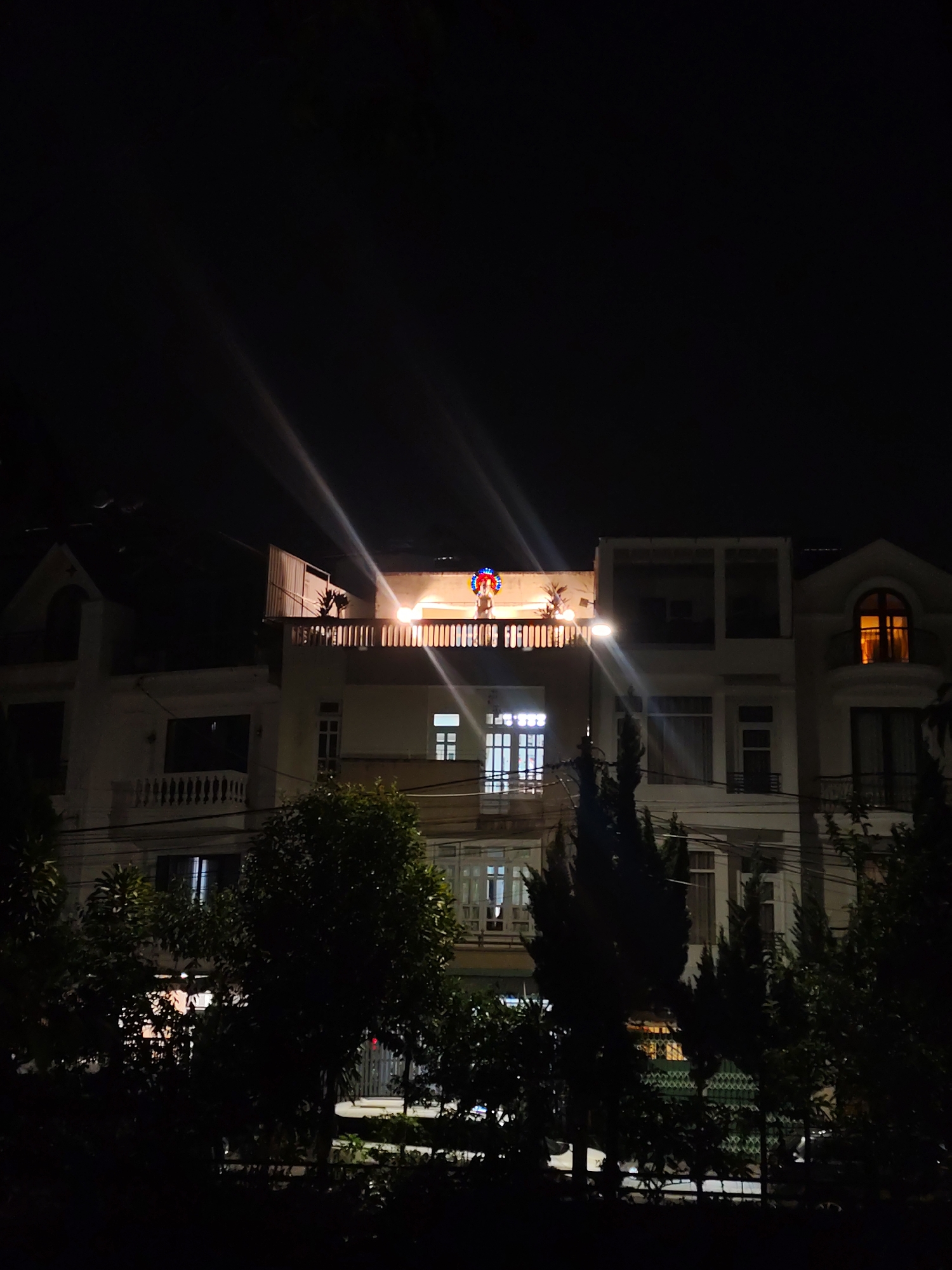 The image captures a nighttime view of several multi-story residential buildings in what appears to be Da Lat, Vietnam, given the location context. The sky is completely dark, illuminated only by artificial lights emanating from the structures.
In the center, a prominent cream-colored building features a large, well-lit rooftop terrace. A powerful light fixture on this terrace casts intense beams of light upwards and outwards, creating significant lens flares that streak across the dark sky. Above this light, a colorful, semi-circular decorative element, possibly illuminated string lights in red, blue, and white, adds a festive touch. Potted plants are also visible on the terrace. Below this, windows on the central building display varying internal illumination, some with blue-toned light and others with a softer white glow.
To the left, another cream-colored building with a distinctive gabled roof section and balconies with white balustrades is partially visible. On the right, a similar building features an arched window glowing with a warm, orange light from within. Dark silhouettes of trees and dense foliage occupy the foreground and mid-ground, obscuring the lower portions of the buildings and adding depth to the scene. Faint power lines crisscross the view.
The overall impression is one of a quiet residential evening, with the bright lights of the central terrace and the colorful decoration suggesting either a special occasion or simply a well-lit home. No explicit human activity is visible, nor is any text discernable in the image.