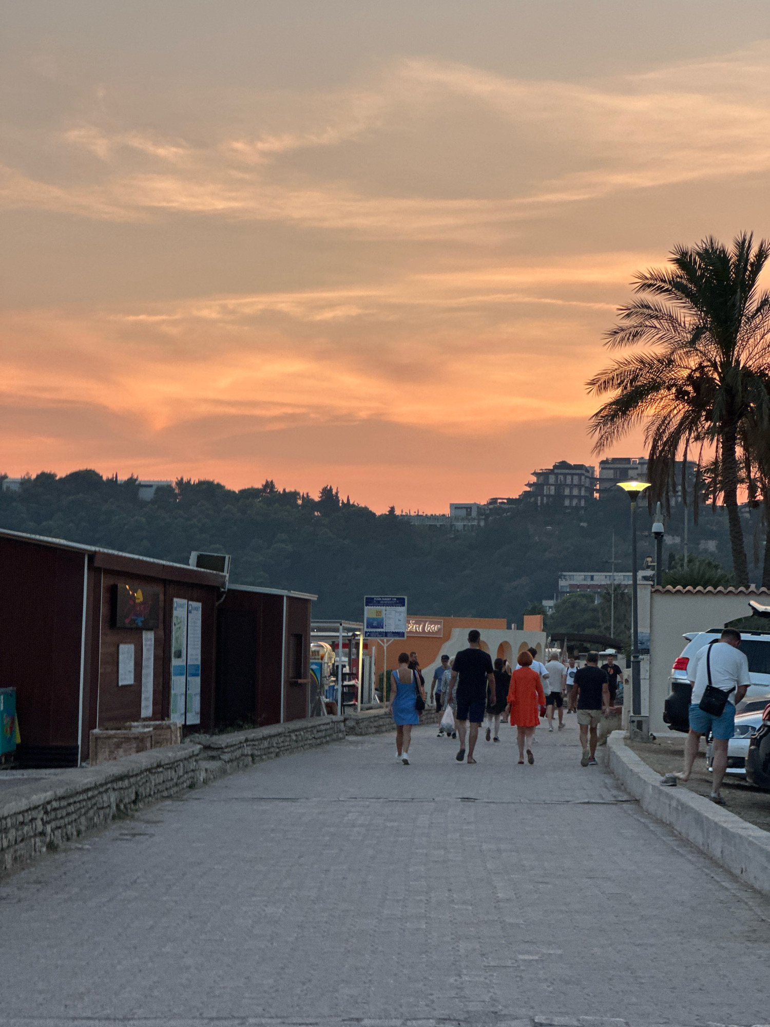 The image depicts a coastal street scene in Boreti, Montenegro, during sunset. A warm, orange-hued sky with thin clouds dominates the upper portion of the frame. A dense, tree-covered hill rises in the background, with several modern buildings nestled on its slopes. A palm tree stands silhouetted against the sky on the right side.
In the foreground, a paved walkway stretches into the distance. On the left side of the walkway are several low-rise structures with dark brown walls and what appear to be commercial establishments. A stone wall runs along the edge of the walkway, separating it from the buildings.
A group of people are walking away from the camera down the paved street. They are casually dressed in summer attire. Among them, a woman in a blue dress, a man in a black t-shirt and dark shorts, a woman in an orange dress, and a man in a white t-shirt and blue shorts are clearly visible. Several parked cars are on the right side of the street, and a lamppost with a bright light is also visible. The overall atmosphere is relaxed and suggests a popular evening promenade.