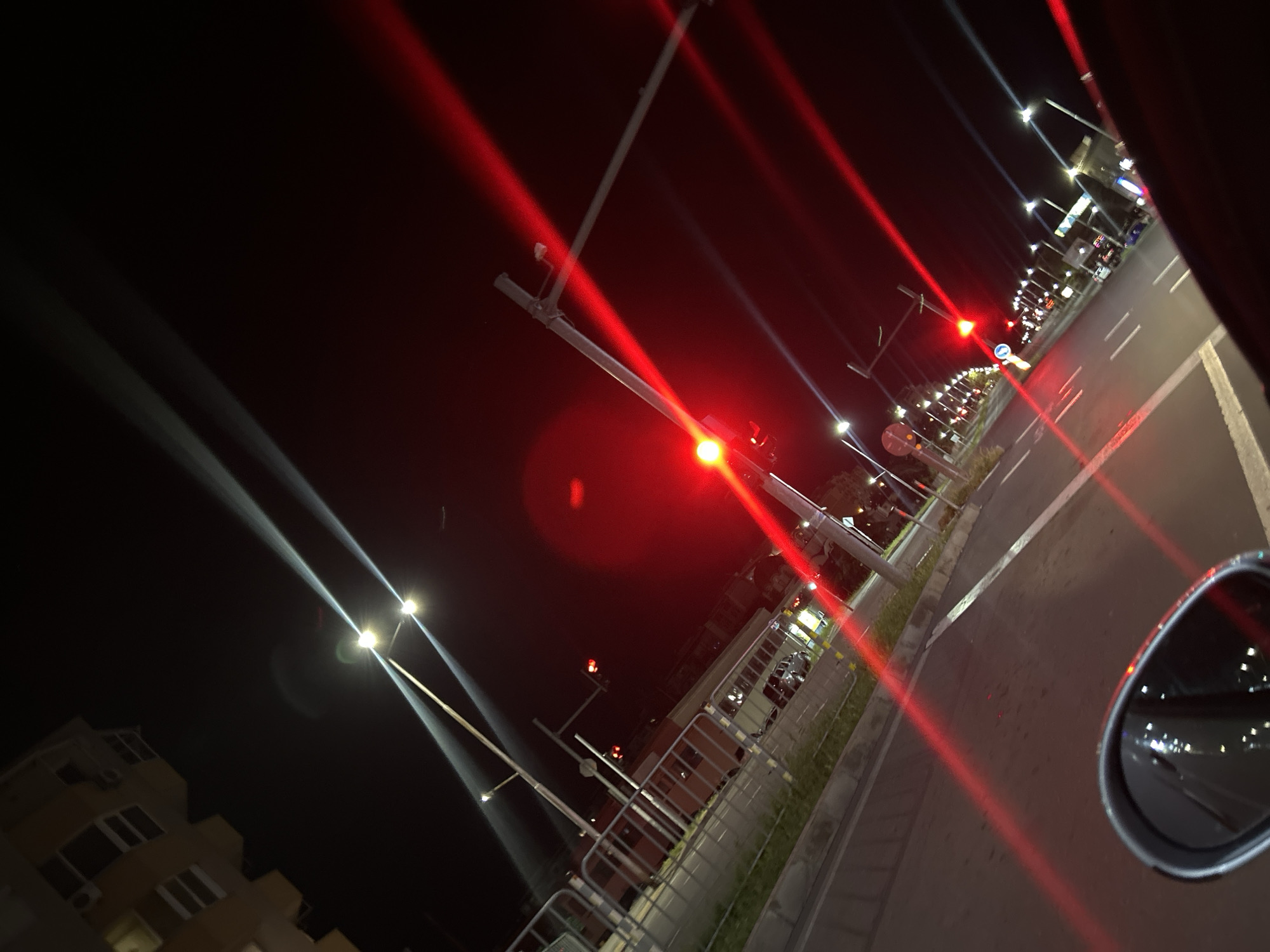 The image depicts a nighttime street scene in an unidentified city. The view is taken from a low angle, likely from a moving vehicle with a side mirror visible in the lower right corner. The street has multiple lanes, with white lines marking the lane divisions. Red streaks emanate from traffic lights along the street, indicating the presence of these lights. Street lamps are visible along the side of the road, casting white light beams. A fence runs parallel to the street. In the lower left corner of the frame, a building is visible, with multiple windows and an air conditioning unit.