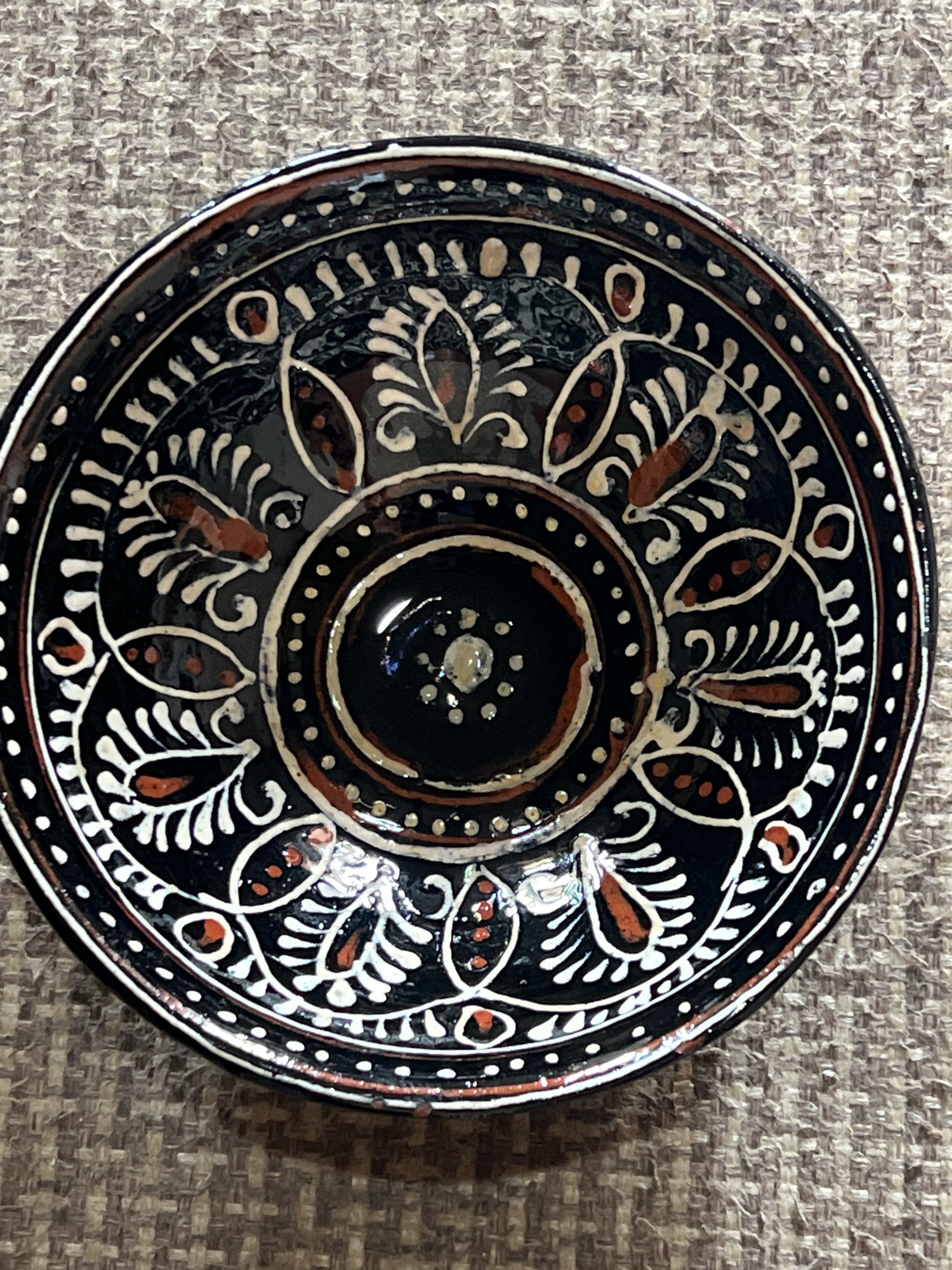 A black ceramic bowl with white and reddish-brown floral and dot patterns rests on a textured brown surface. The bowl is intricately decorated with concentric circles of dots and stylized floral motifs that feature curved lines and pointed shapes. The center of the bowl has a small, shiny indentation. The background appears to be a woven fabric with a mottled brown and gray appearance. The image was taken from a top-down perspective, with the bowl slightly angled. The location is not specified in the image or metadata.