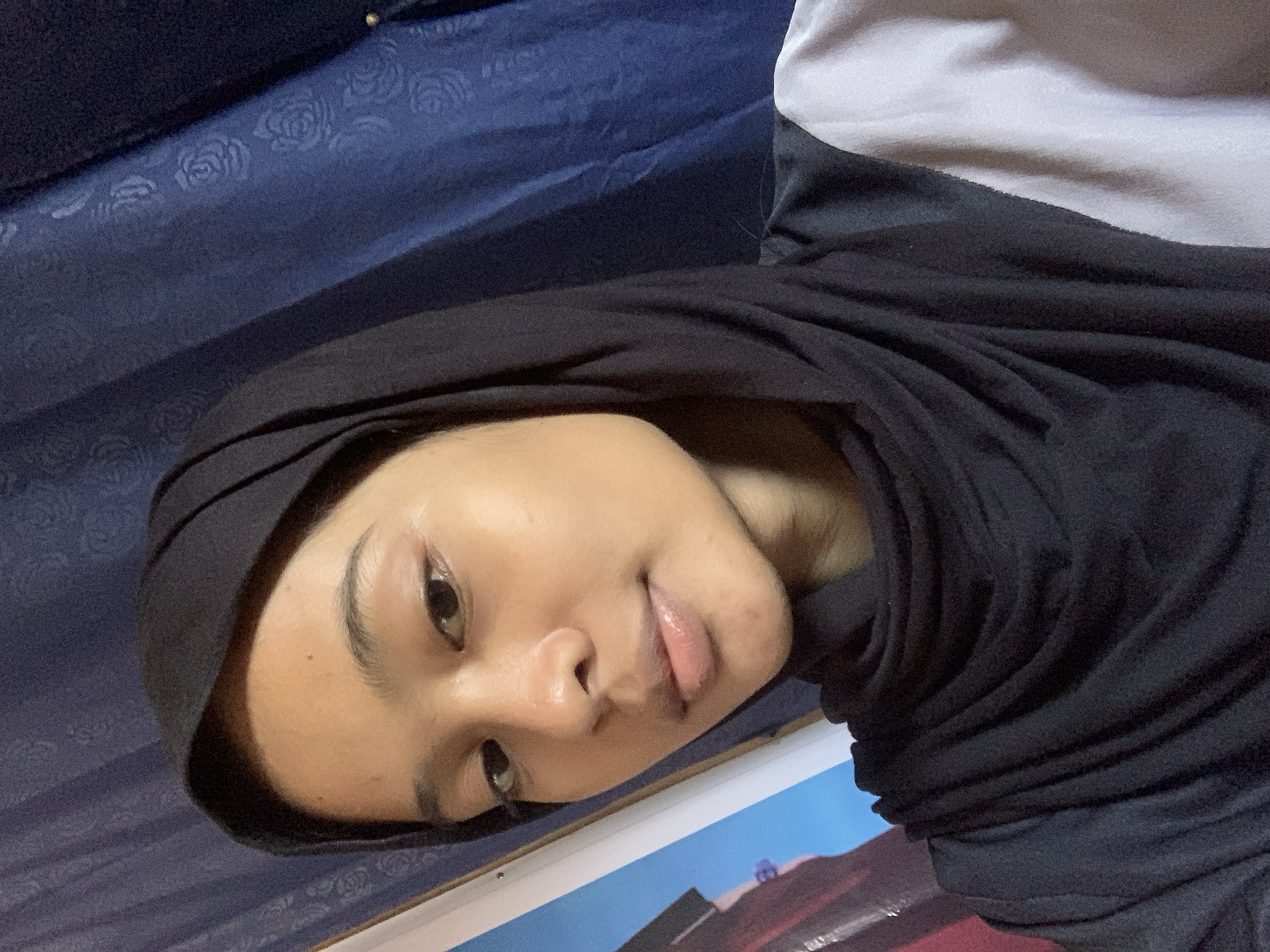 The image is a close-up, selfie-style portrait of a young woman wearing a black hijab. She is looking directly at the camera with her face centered. The background consists of a blue, textured curtain with a subtle floral pattern. To the left of the woman, a portion of a painting or poster with blue and red tones is visible. The woman's clothing appears to be a black top with a lighter grey or white section on the right side. The lighting suggests an indoor setting.
