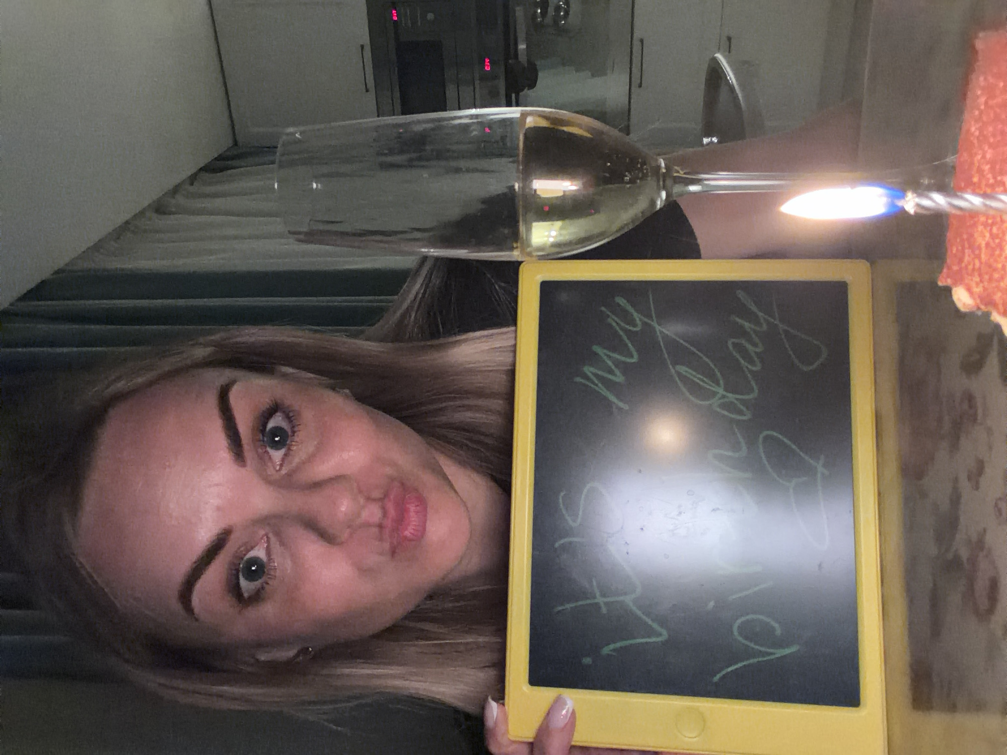 A woman with blonde hair holds a yellow tablet with "It's my birthday" written on it. A wine glass filled with a light-colored liquid is positioned above the tablet. A birthday cake with a lit candle is visible in the upper right corner. The setting appears to be indoors, with cabinets and curtains visible in the background. There is no definitive evidence in the image to confirm the location is Votkinsk, Russia.